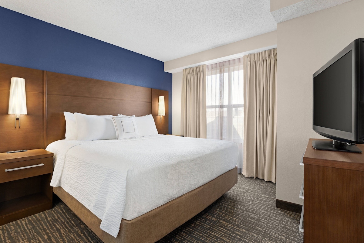 A hotel room with a large bed, a TV on a wooden stand, a nightstand with a lamp, and a window with curtains.