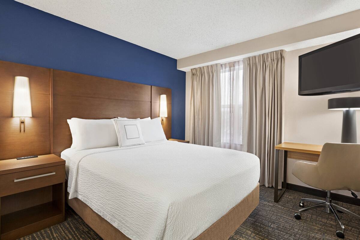 A hotel room with a large bed, a desk, a chair, a TV, and a nightstand.