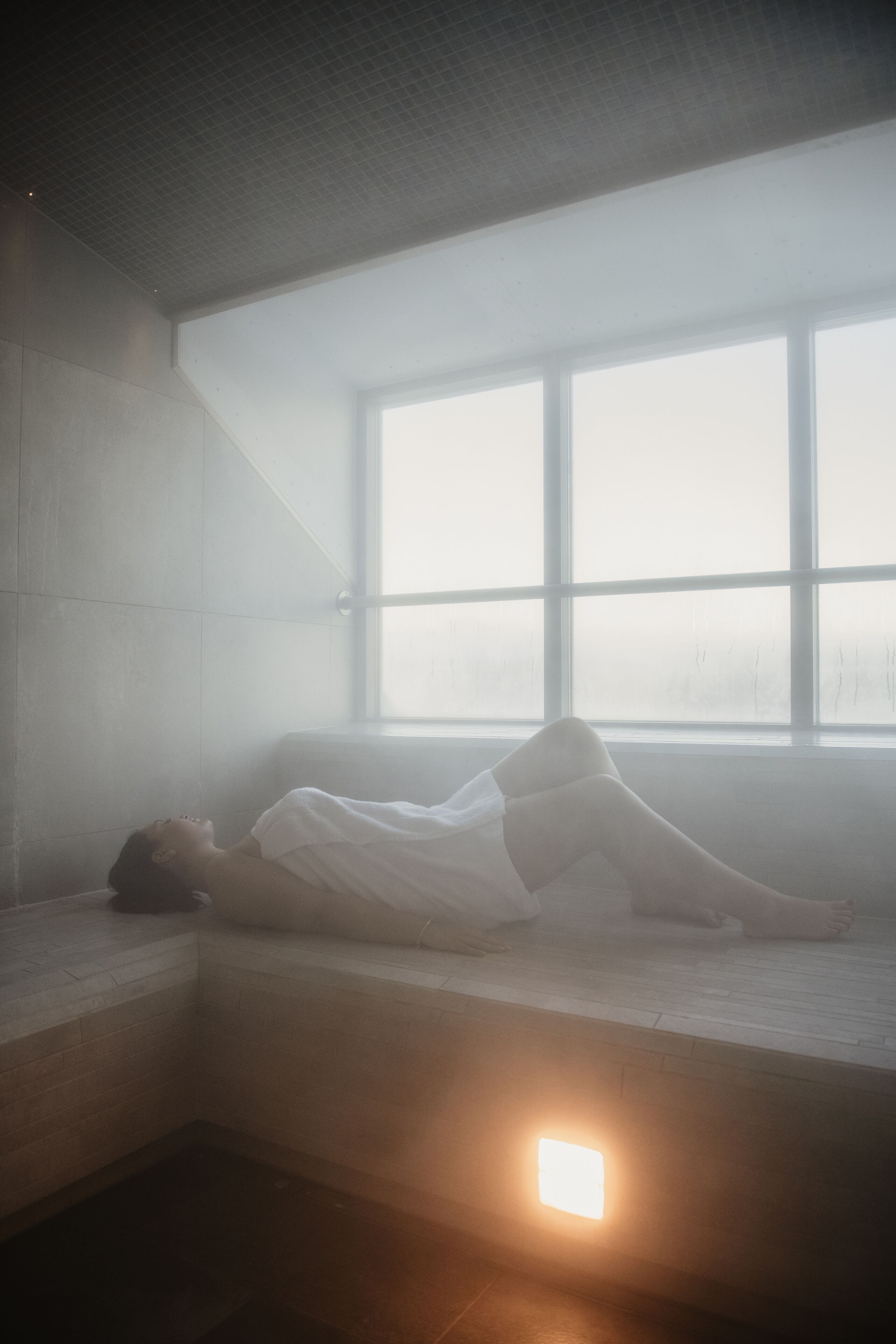 Una persona recostada en un banco dentro de una habitación con un ventanal grande, un muro de azulejos y una fuente de luz en el suelo.