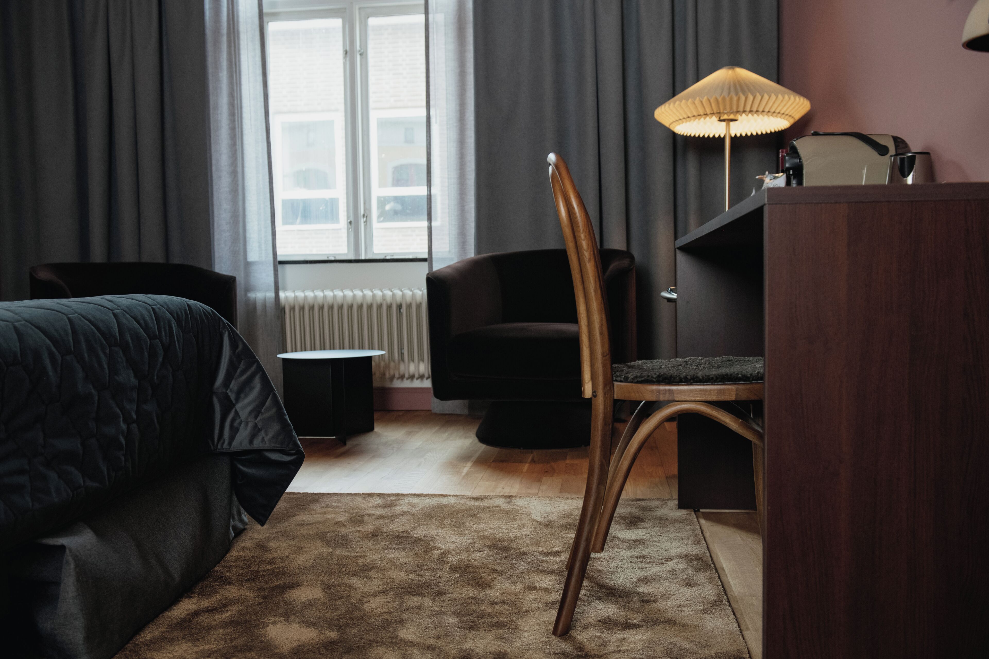 A hotel room with a bed, a desk, a chair, a lamp, and a view of the outside through a window.