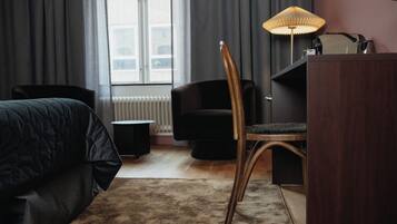 A hotel room with a bed, a desk, a chair, a lamp, and a view of the outside through a window.