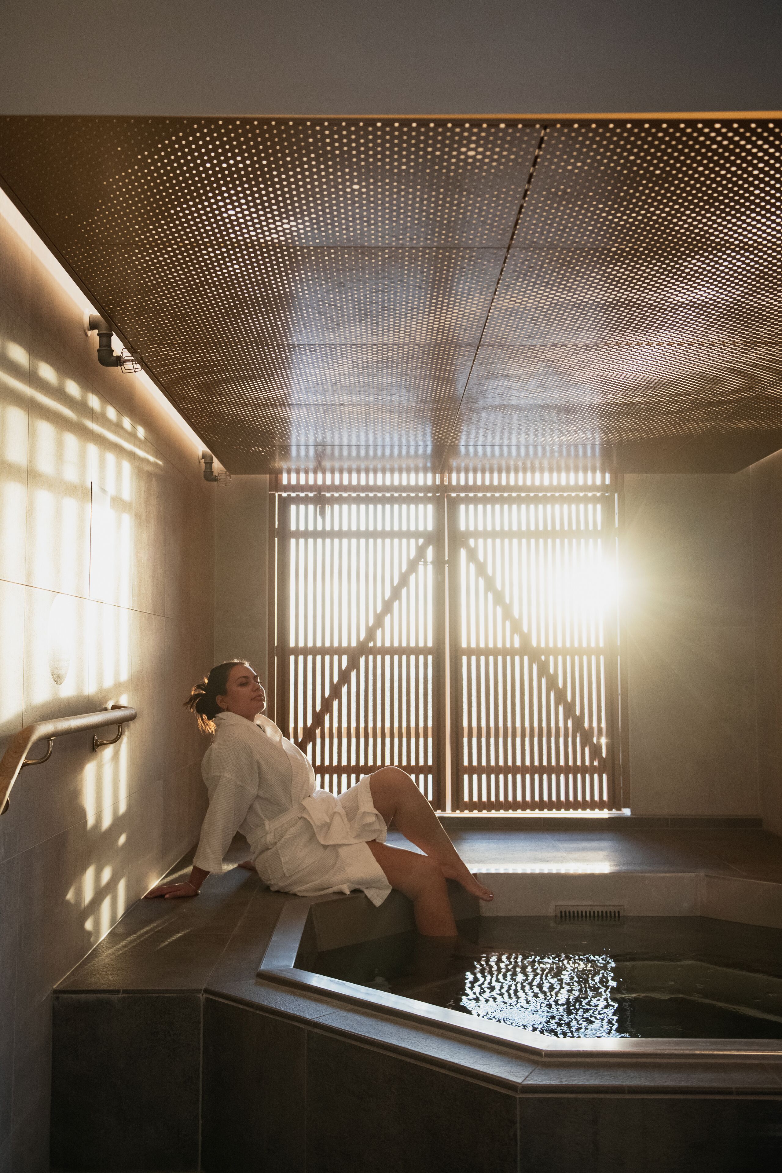 Una persona relajándose en un jacuzzi con techo de madera y vista a un edificio.