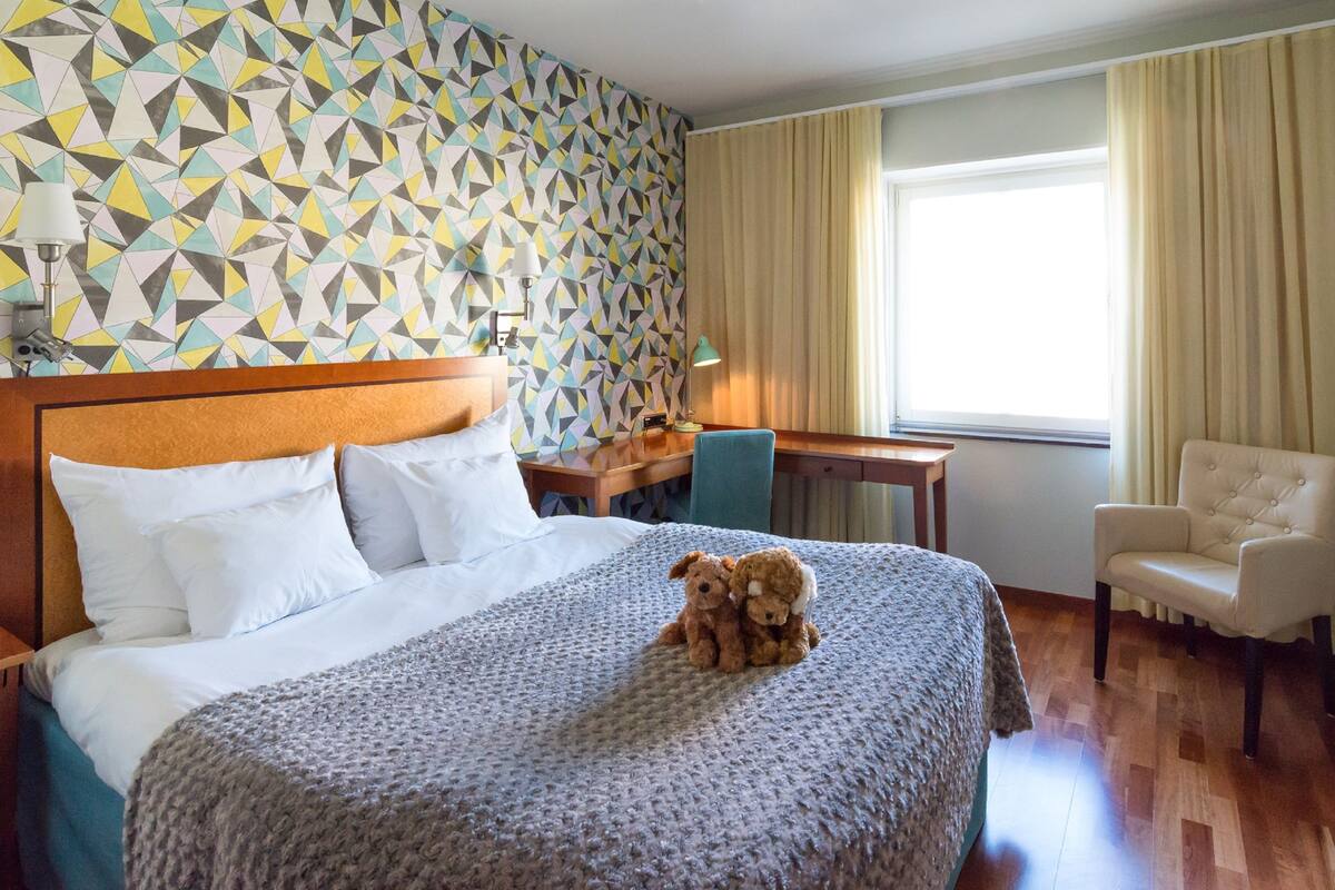 A hotel room with a large bed, a desk, a chair, and a teddy bear on the bed.