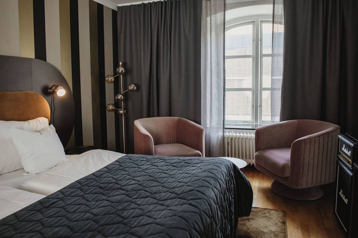 A modern bedroom with a large bed, two armchairs, a floor lamp, and a view of the city through a window.