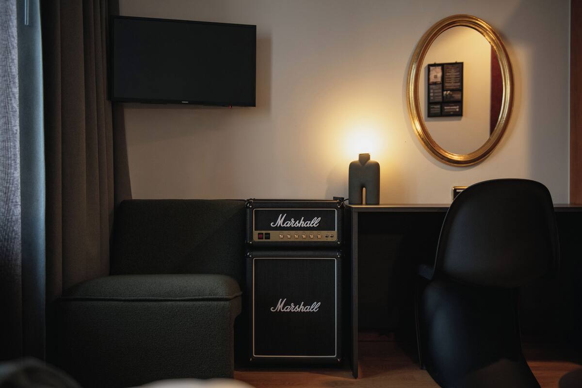 A hotel room with a dark grey sofa, a Marshall amplifier, a flat-screen TV mounted on the wall, and a mirror with a gold frame.