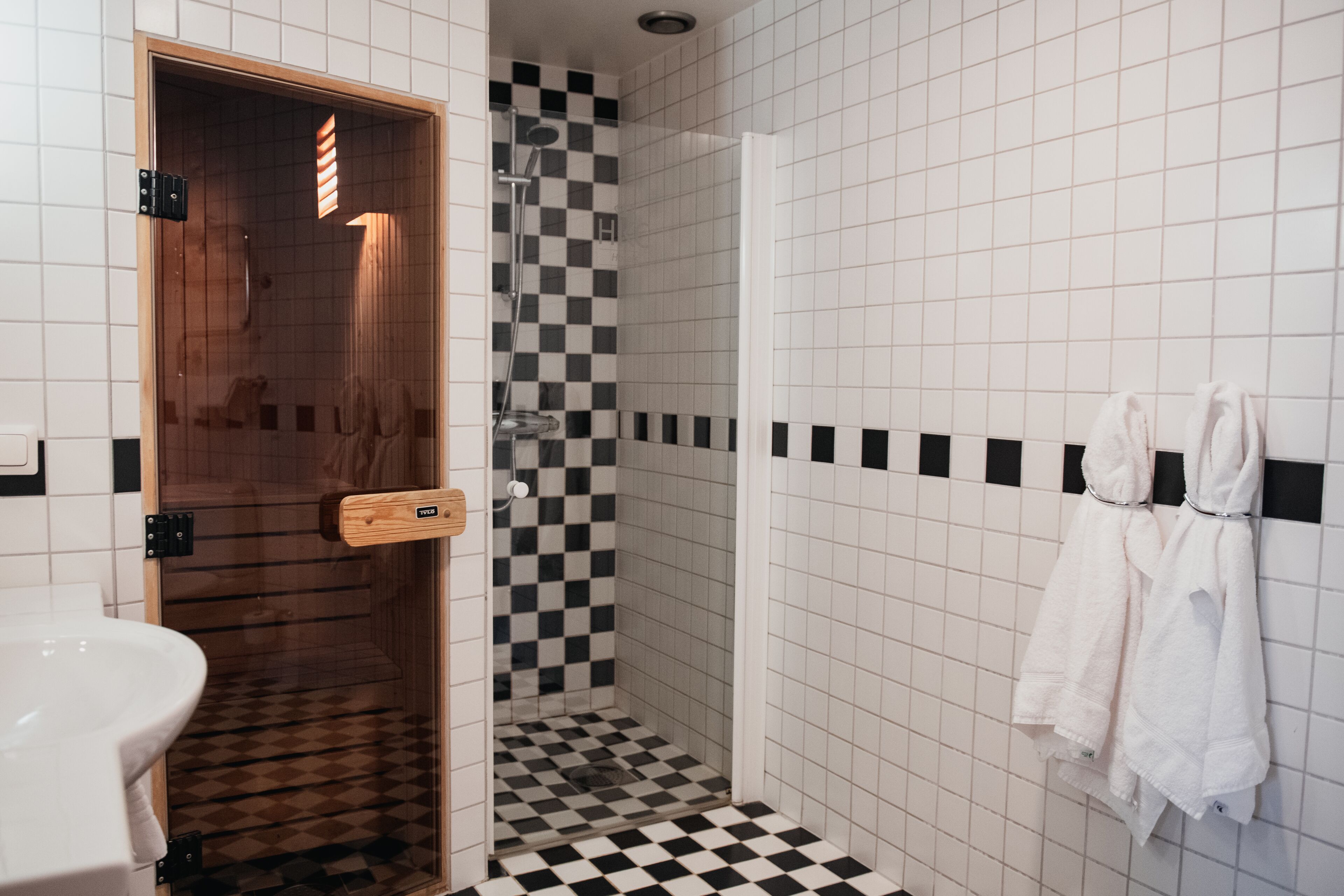 A bathroom with a checkered floor, a wooden sauna door, a white sink, and towels hanging on the wall.