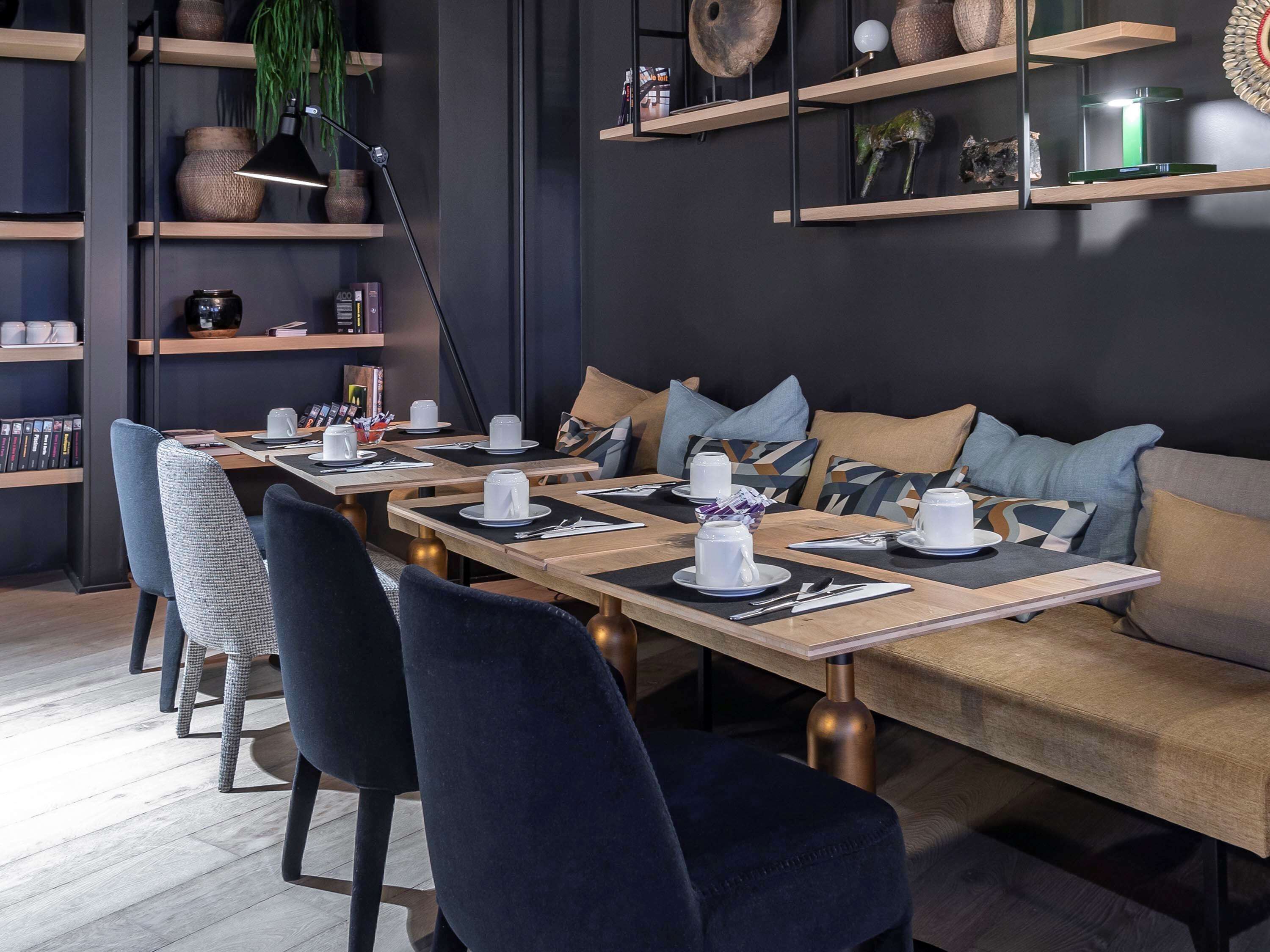 A modern dining area with wooden tables and chairs, a sofa with cushions, and shelves with decorative items.