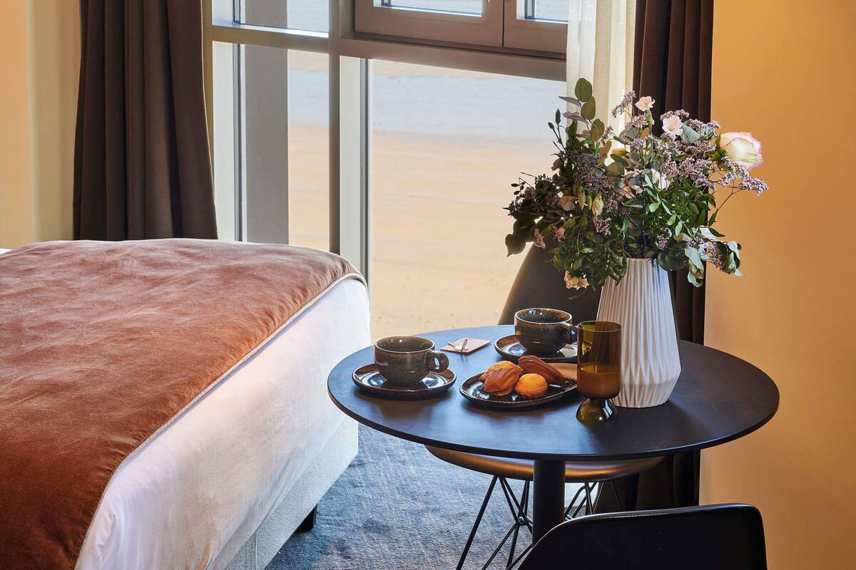 A hotel room with a bed, a table with a vase of flowers, a plate of pastries, and a cup.