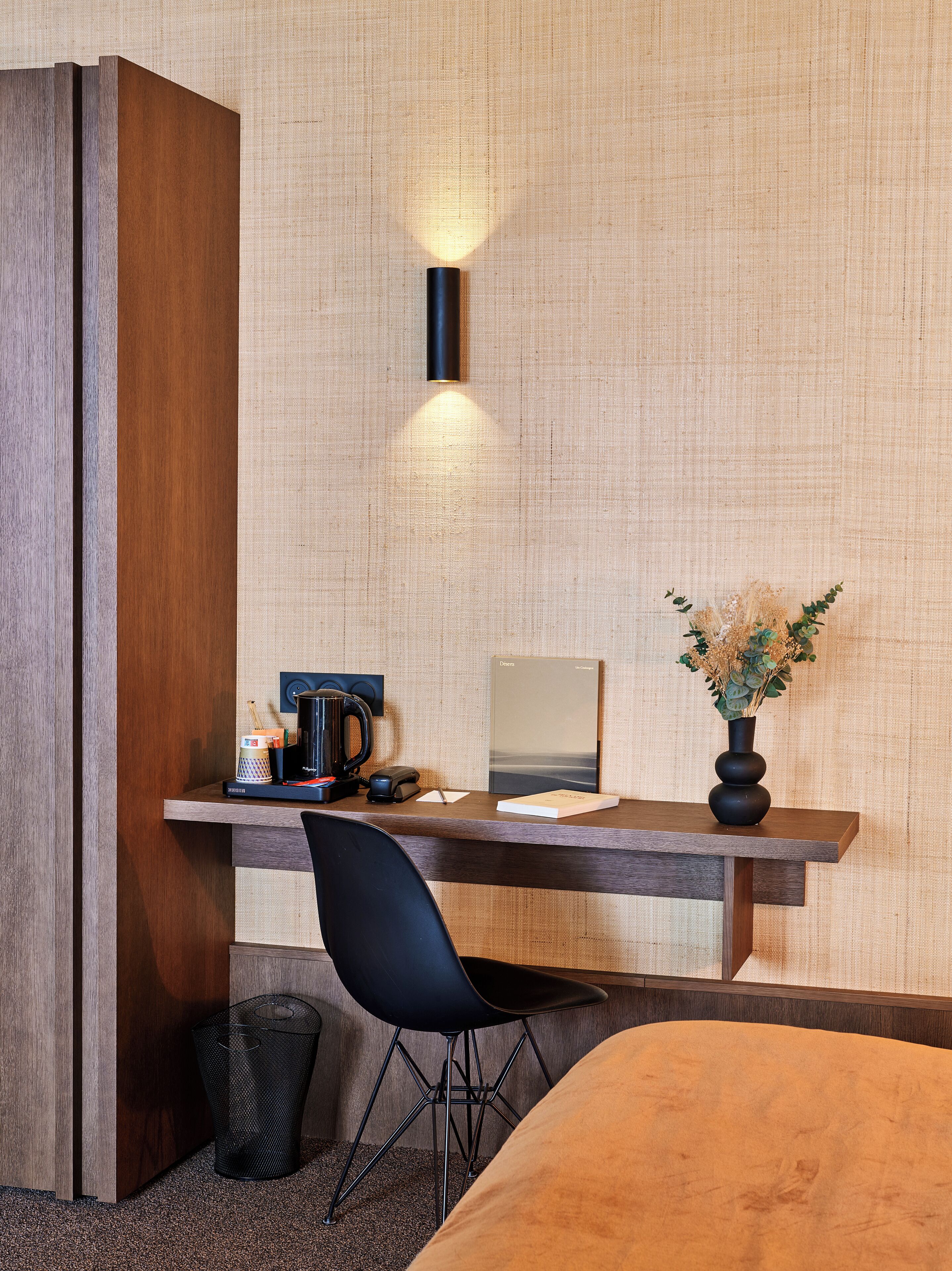 A hotel room with a wooden desk, a black chair, a small trash bin, and a vase with dried flowers.