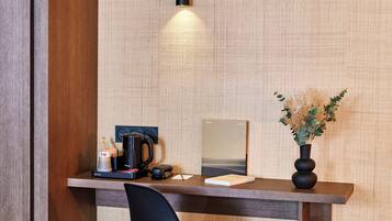 A hotel room with a wooden desk, a black chair, a small trash bin, and a vase with dried flowers.