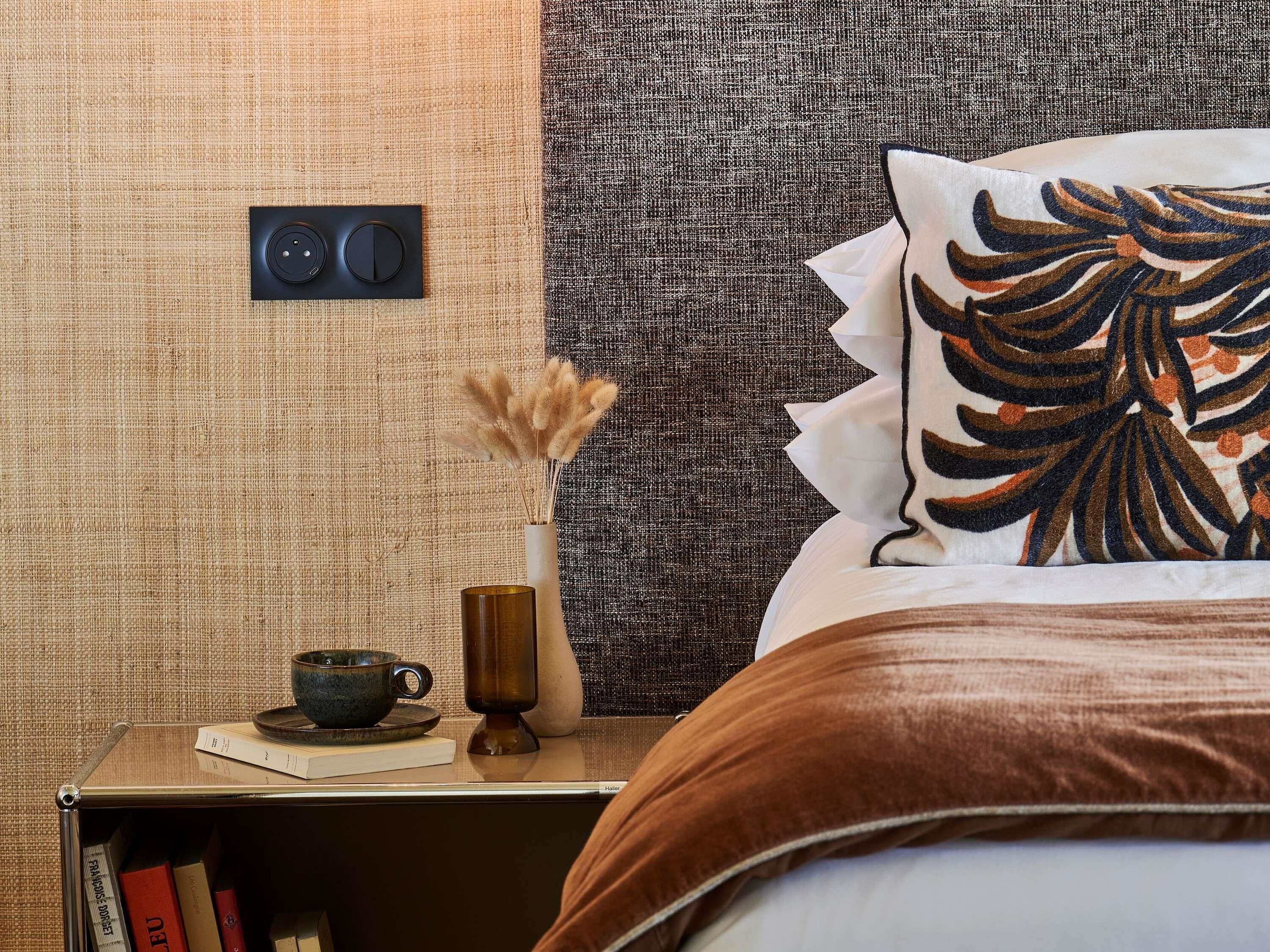 Quarto de hotel bem arrumado com uma cama, mesa de cabeceira com uma xícara, livros e um vaso com flores secas.