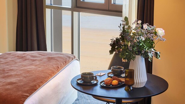A hotel room with a bed, a table with a vase of flowers, a cup, and a plate of pastries. - Mercure St Malo Front De Mer (Saint-Malo)