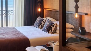 A hotel room with a bed, a bedside table with a cup and a book, a view of the sea through the window, and a wall-mounted light fixture. - Mercure St Malo Front De Mer (Saint-Malo)