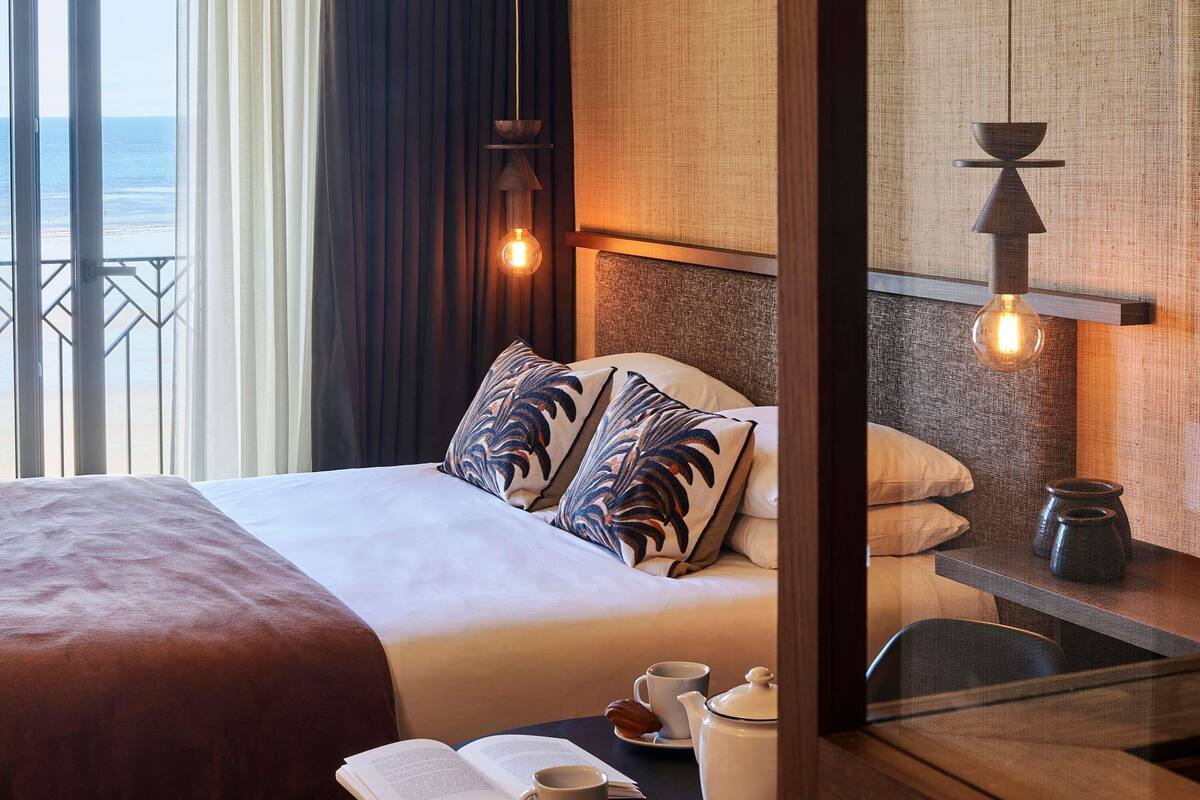 A hotel room with a bed, a bedside table with a cup and a book, a view of the sea through the window, and a wall-mounted light fixture.