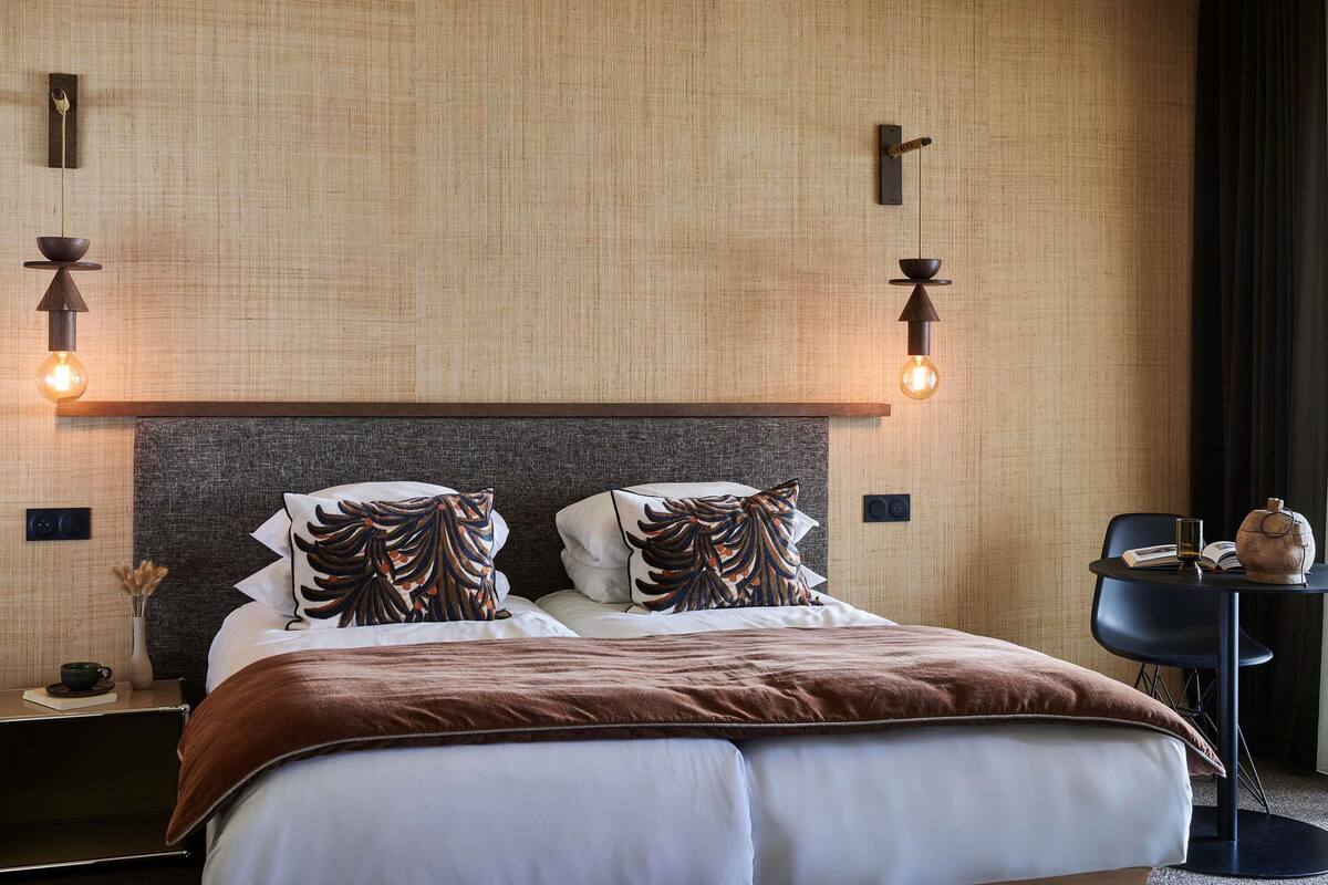 A hotel room with a large bed, two bedside tables, a small table with a chair, and a wall-mounted light fixture.