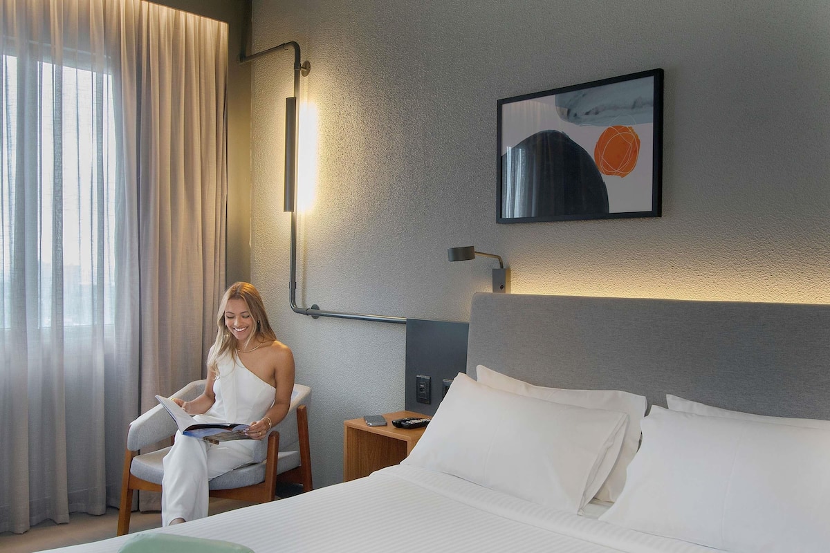 A hotel room with a bed, a chair, a bedside table with a lamp, and a painting on the wall.