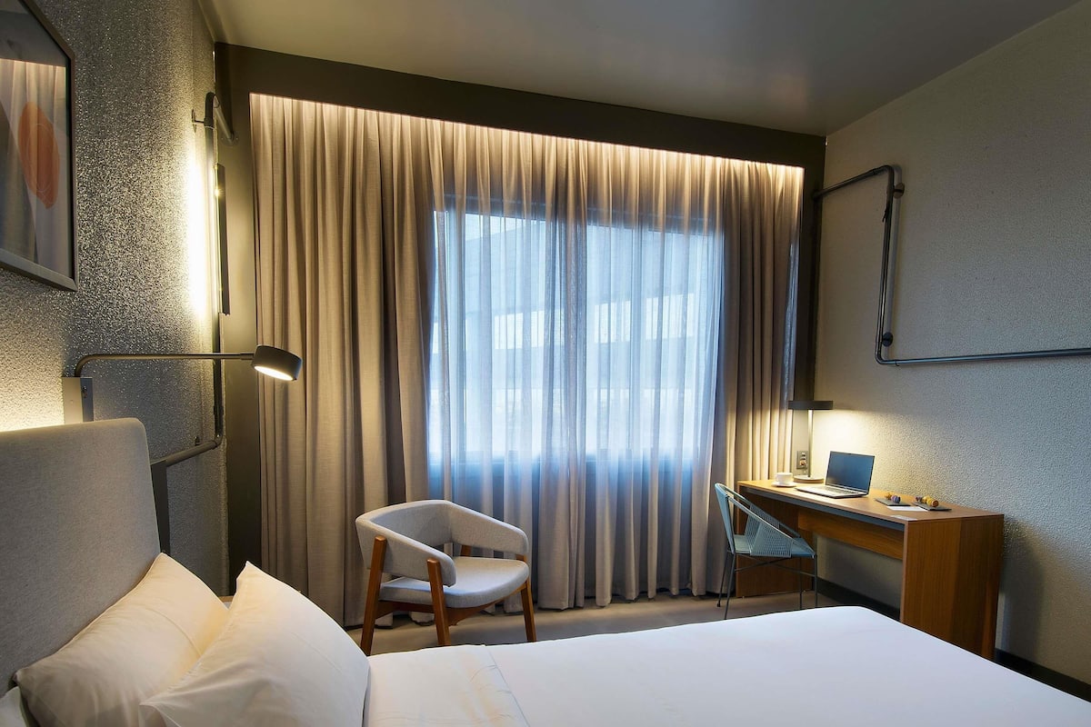 A hotel room with a bed, a desk, a chair, and a large window with curtains.