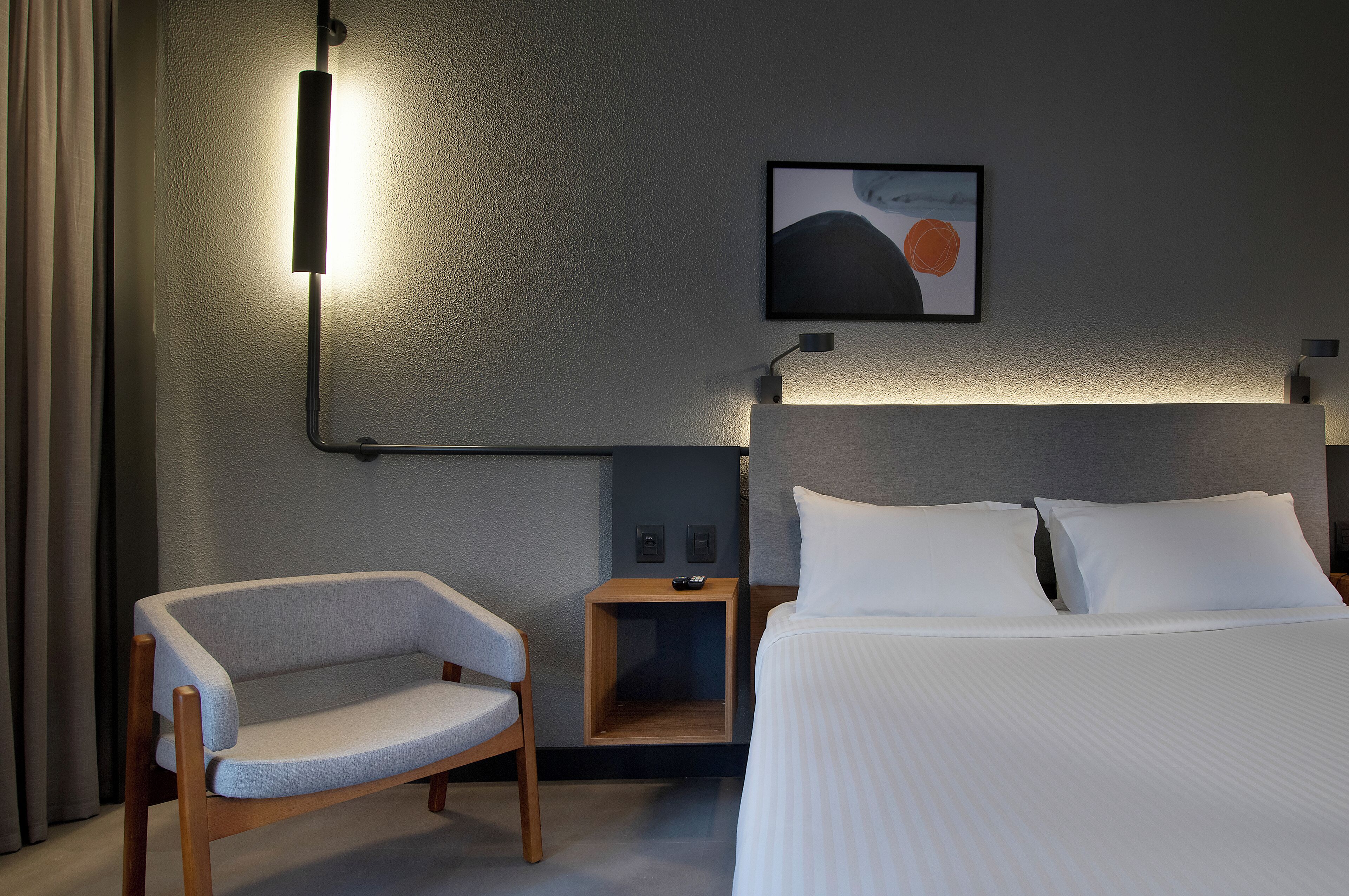 Una habitación de hotel con una cama, una silla, una mesita de noche y una lámpara fijada a la pared.