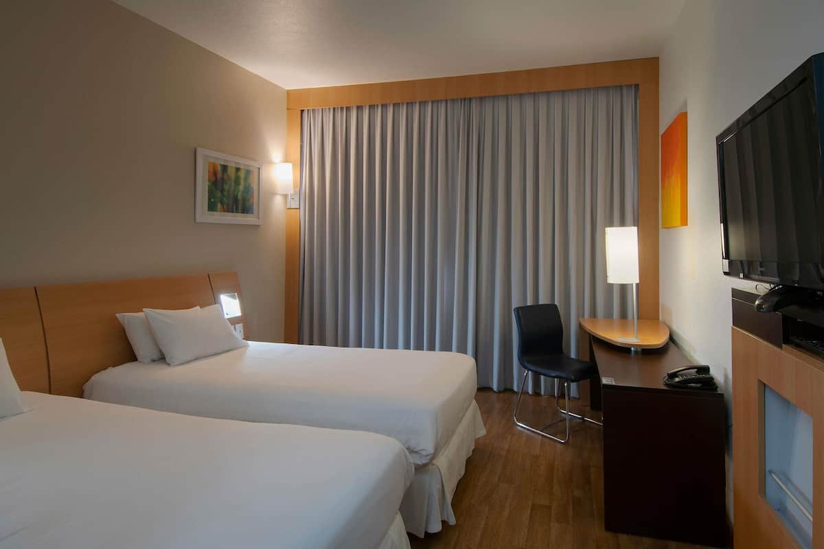 A hotel room with two beds, a desk with a chair, a television, and a large window with curtains.