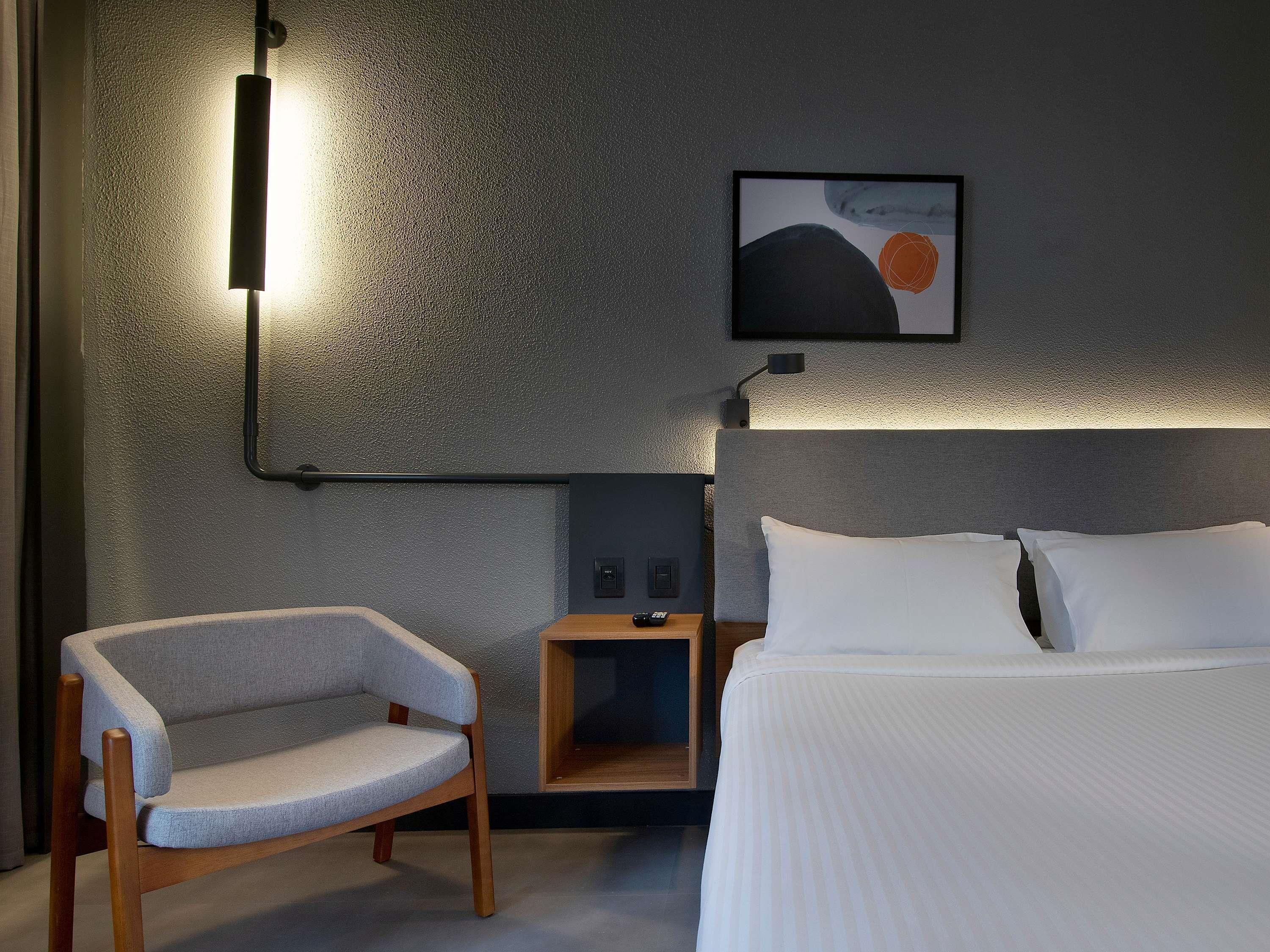 A modern hotel room with a bed, a chair, a nightstand, and a wall-mounted lamp.