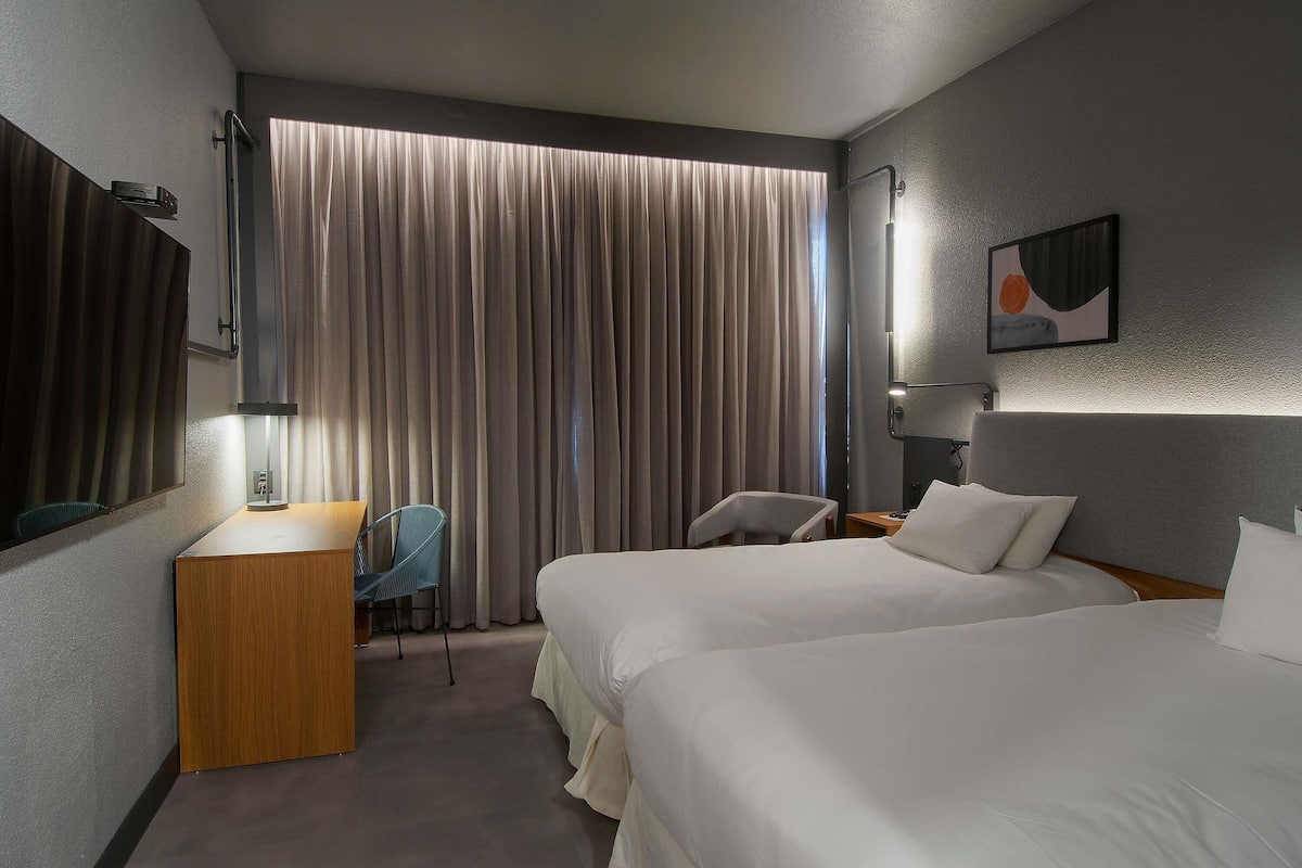A hotel room with two beds, a desk, a chair, a large window with curtains, and a painting on the wall.