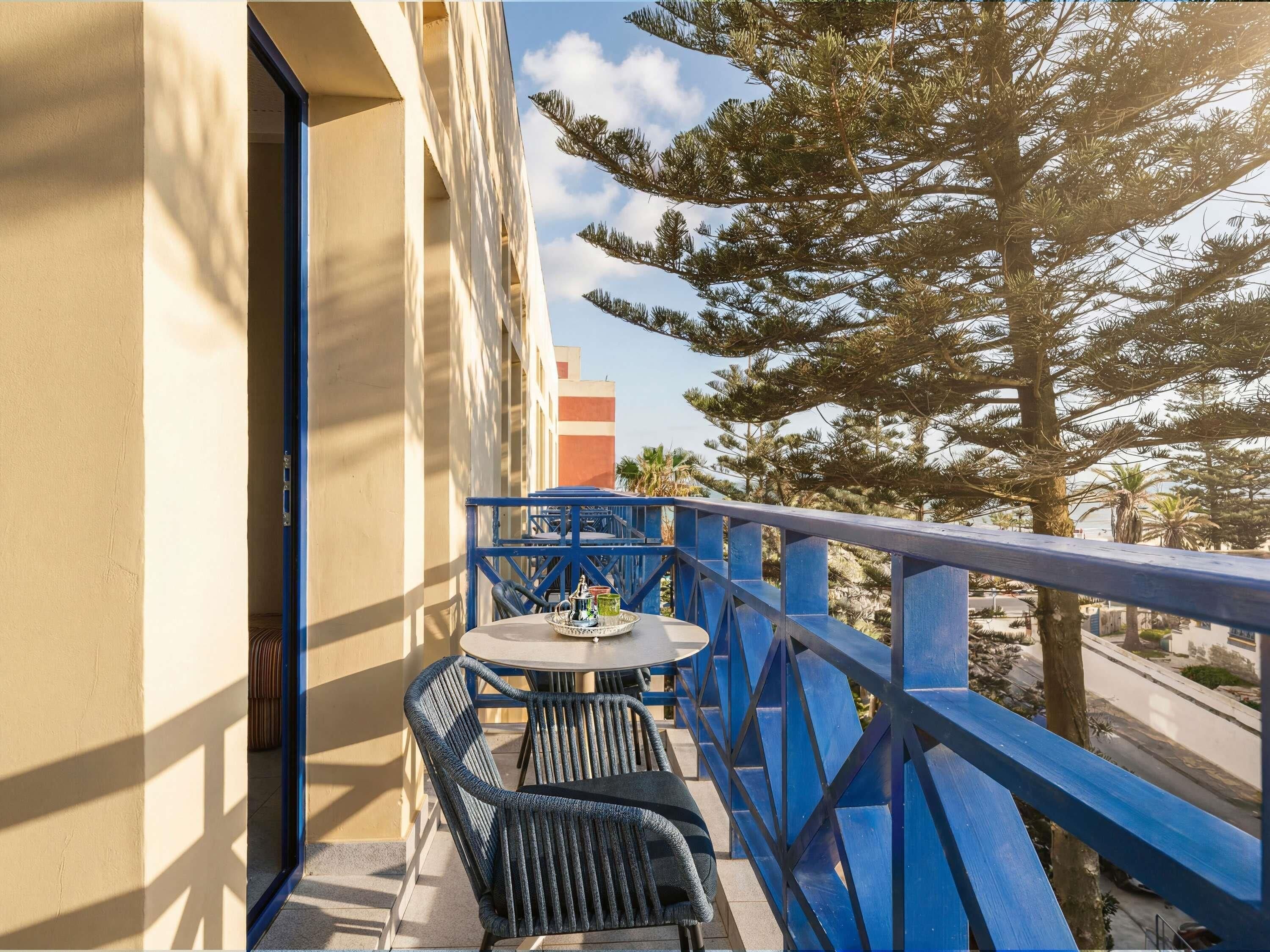 A balcony with a table and chairs, a blue railing, and a view of trees and buildings.