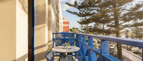 A balcony with a table and chairs, a blue railing, and a view of trees and buildings.
