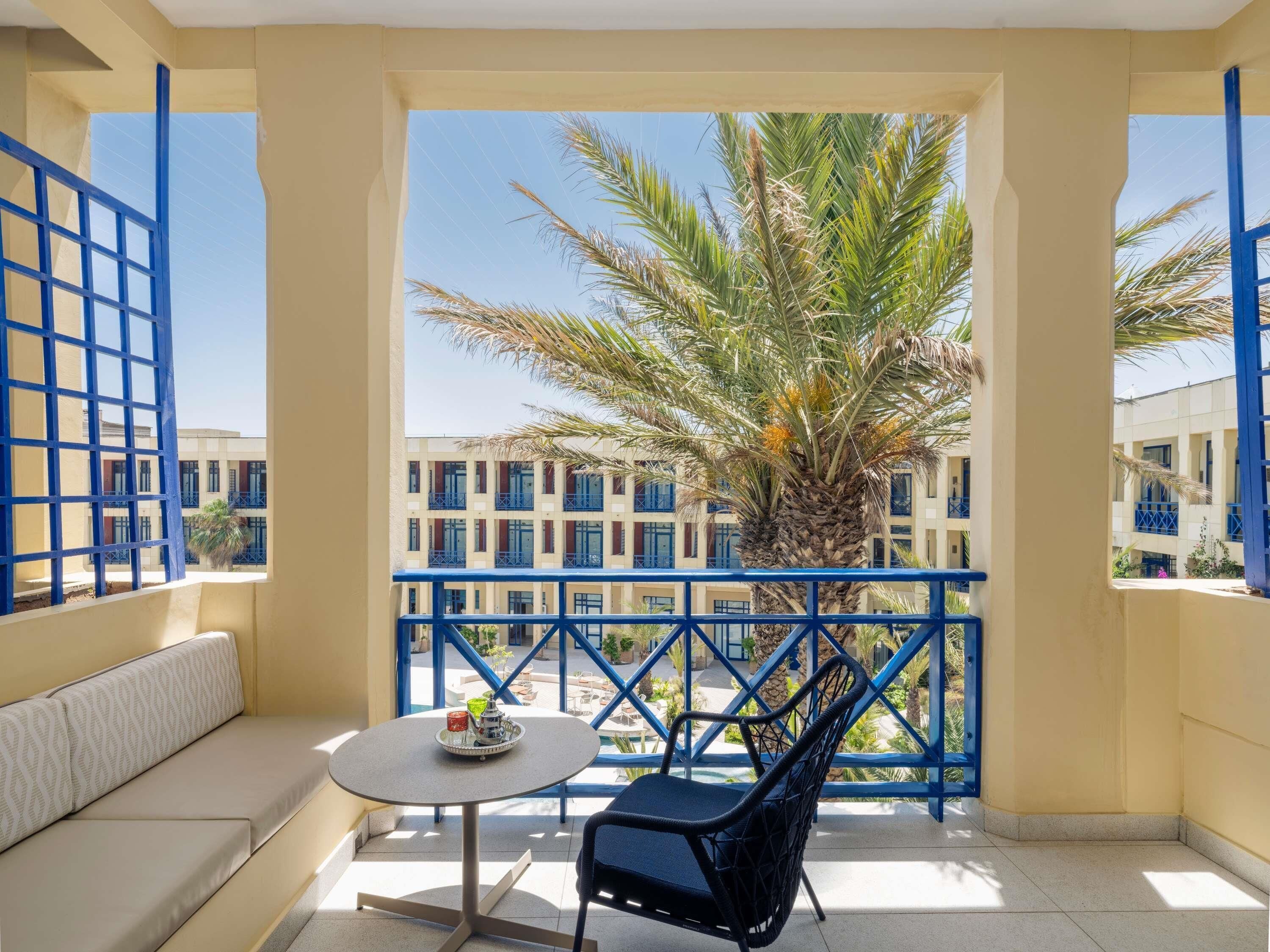 Un balcón con sofá, mesa y silla, con vistas a un edificio de balcones azules y palmeras.