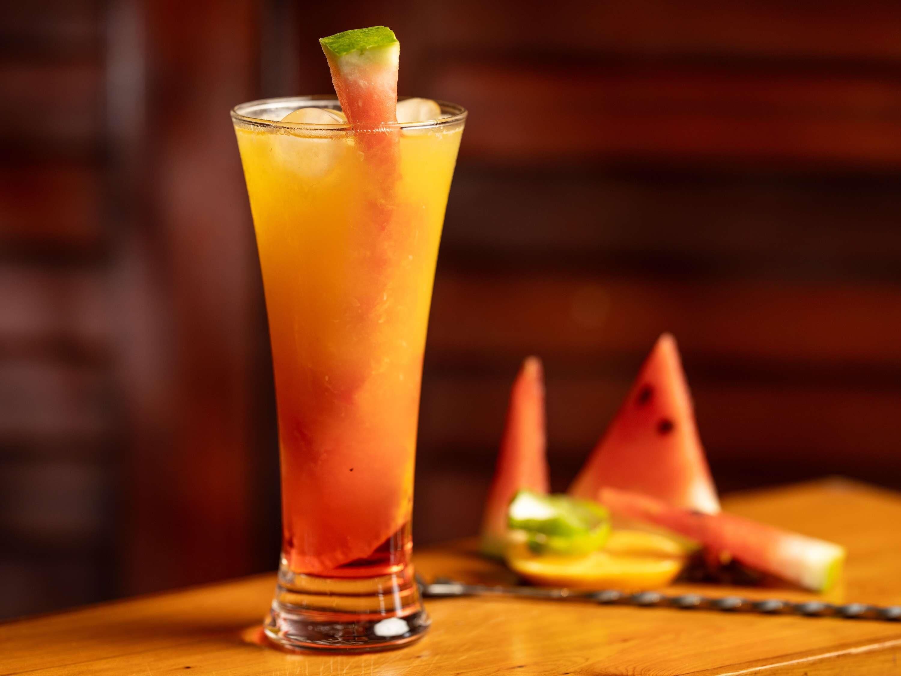 A tall glass with a layered drink, a watermelon slice, and a lime wedge on a wooden table.