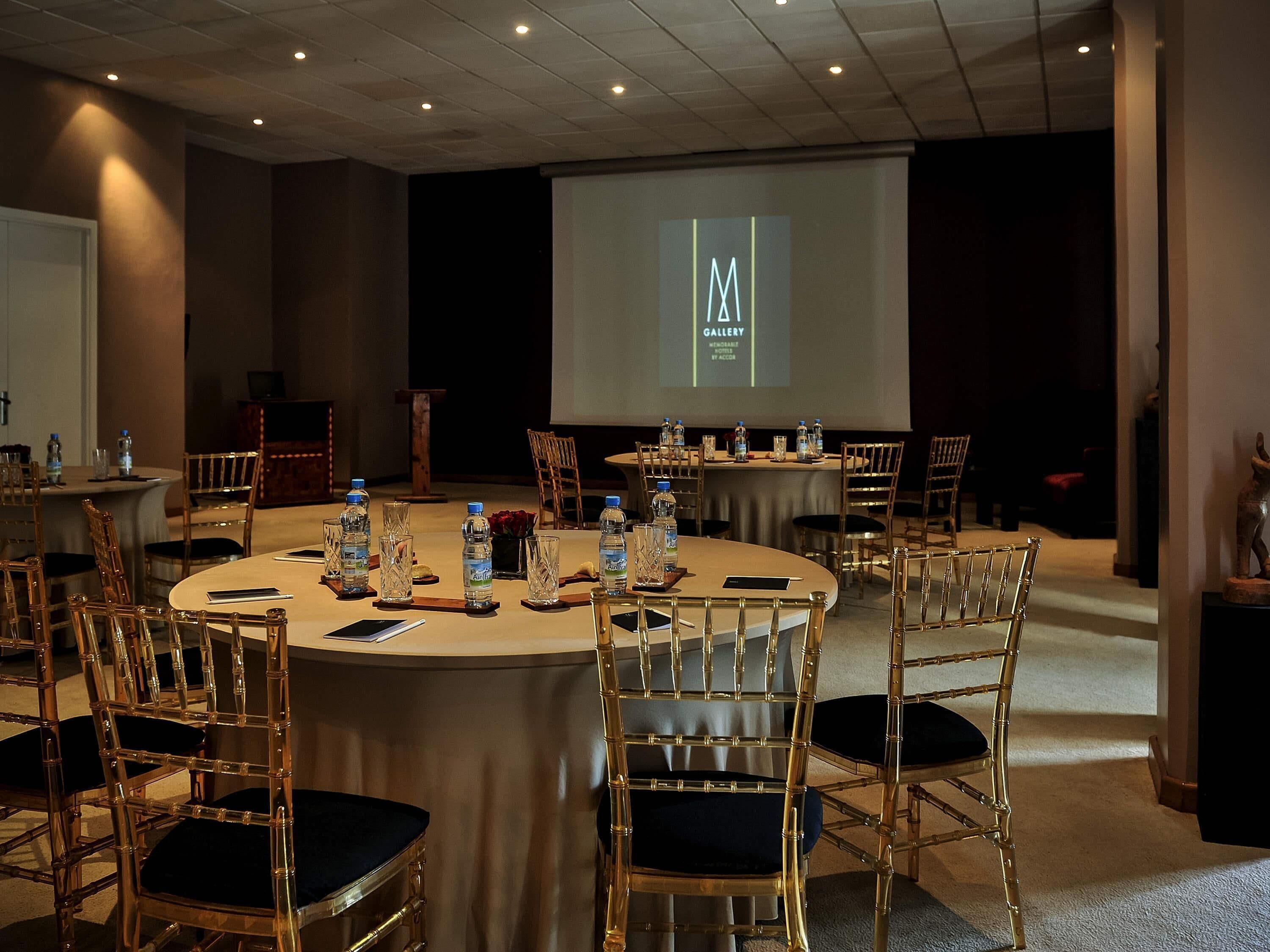 Un salón de conferencias con mesas redondas, sillas, botellas de agua y una pantalla de proyector mostrando un logotipo.