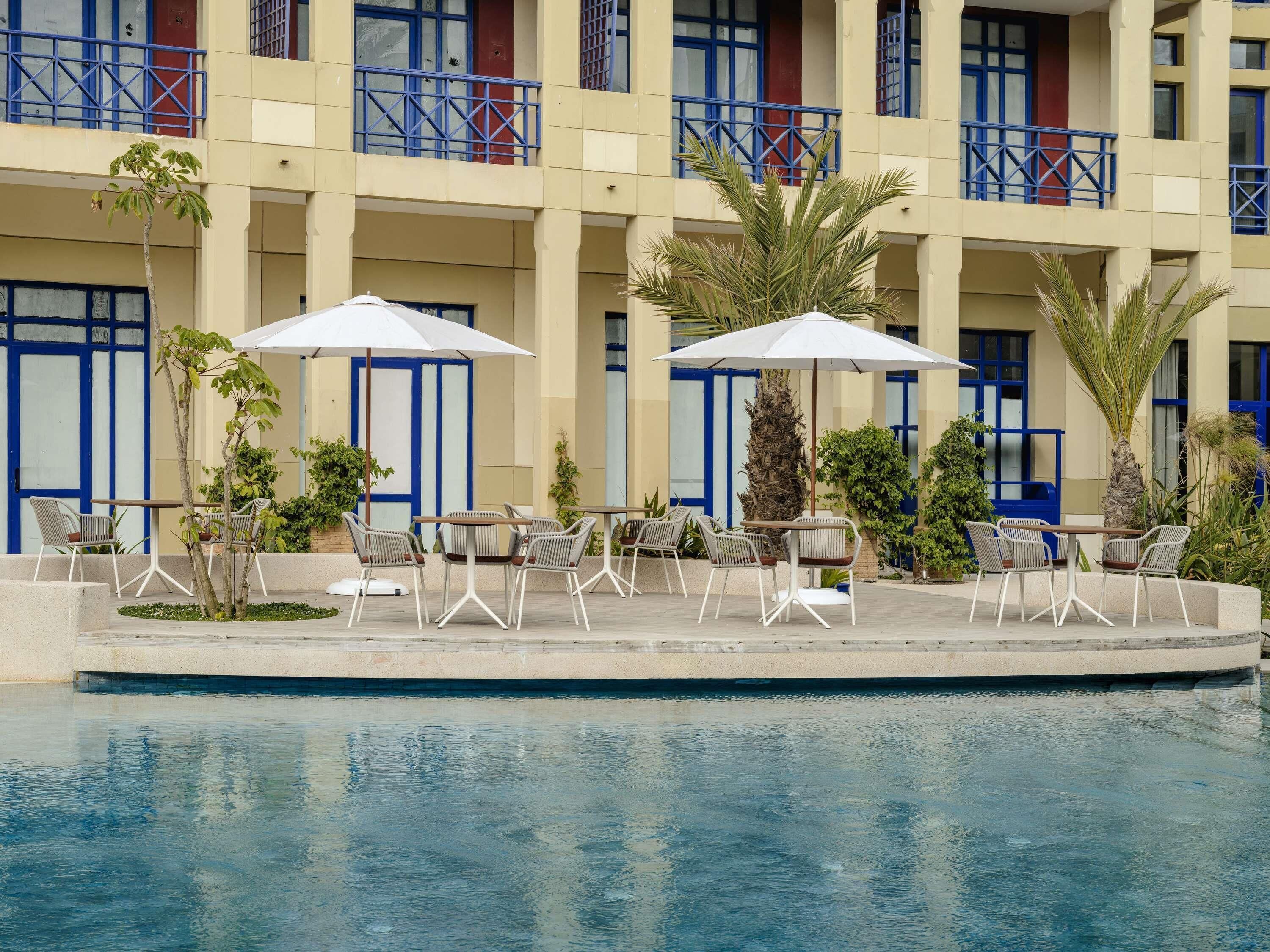 A poolside area with outdoor seating and umbrellas, a building with blue-framed windows, and a palm tree.