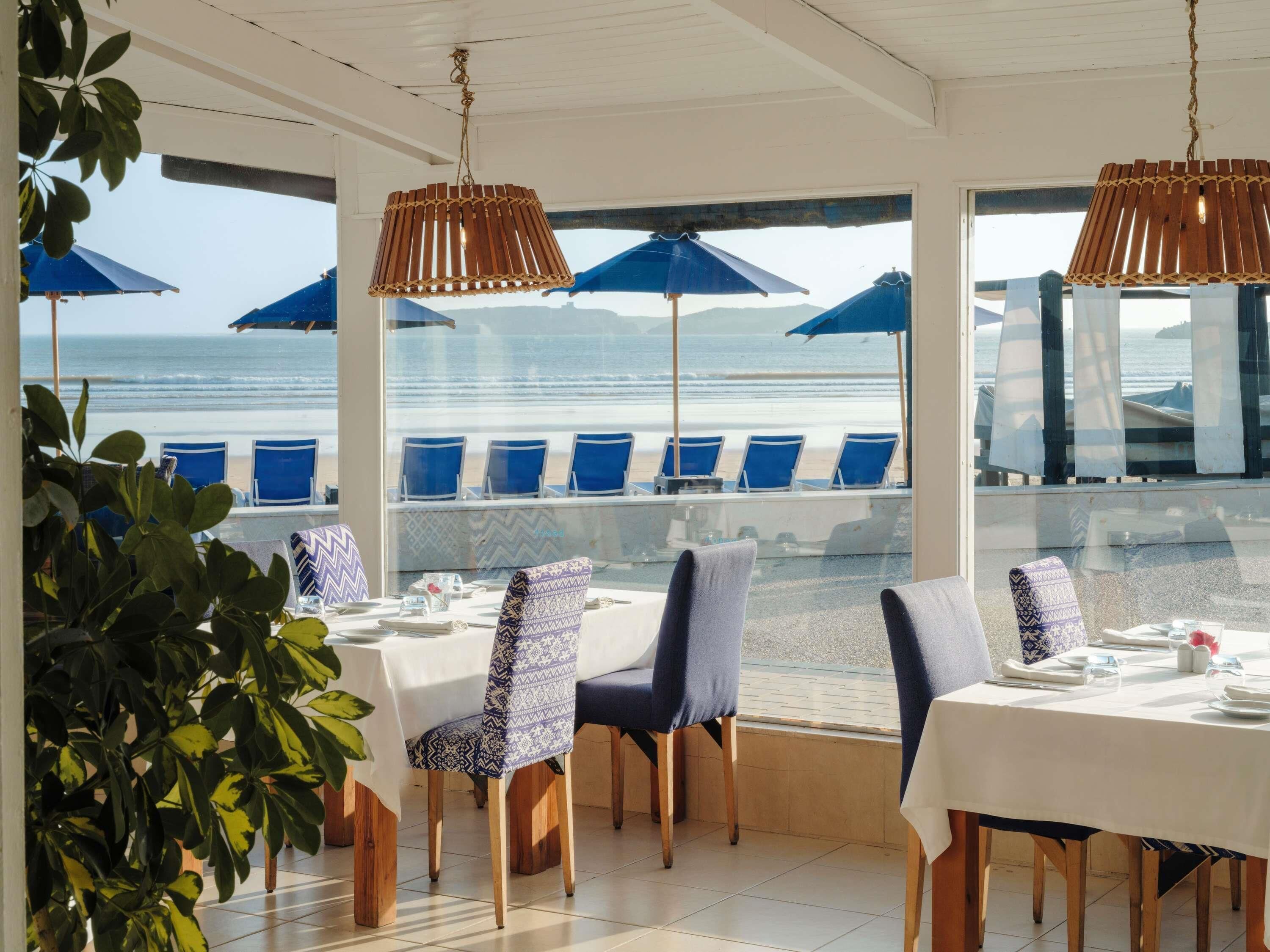 A restaurant with a view of the beach, featuring blue chairs, white tables, and hanging lamps.