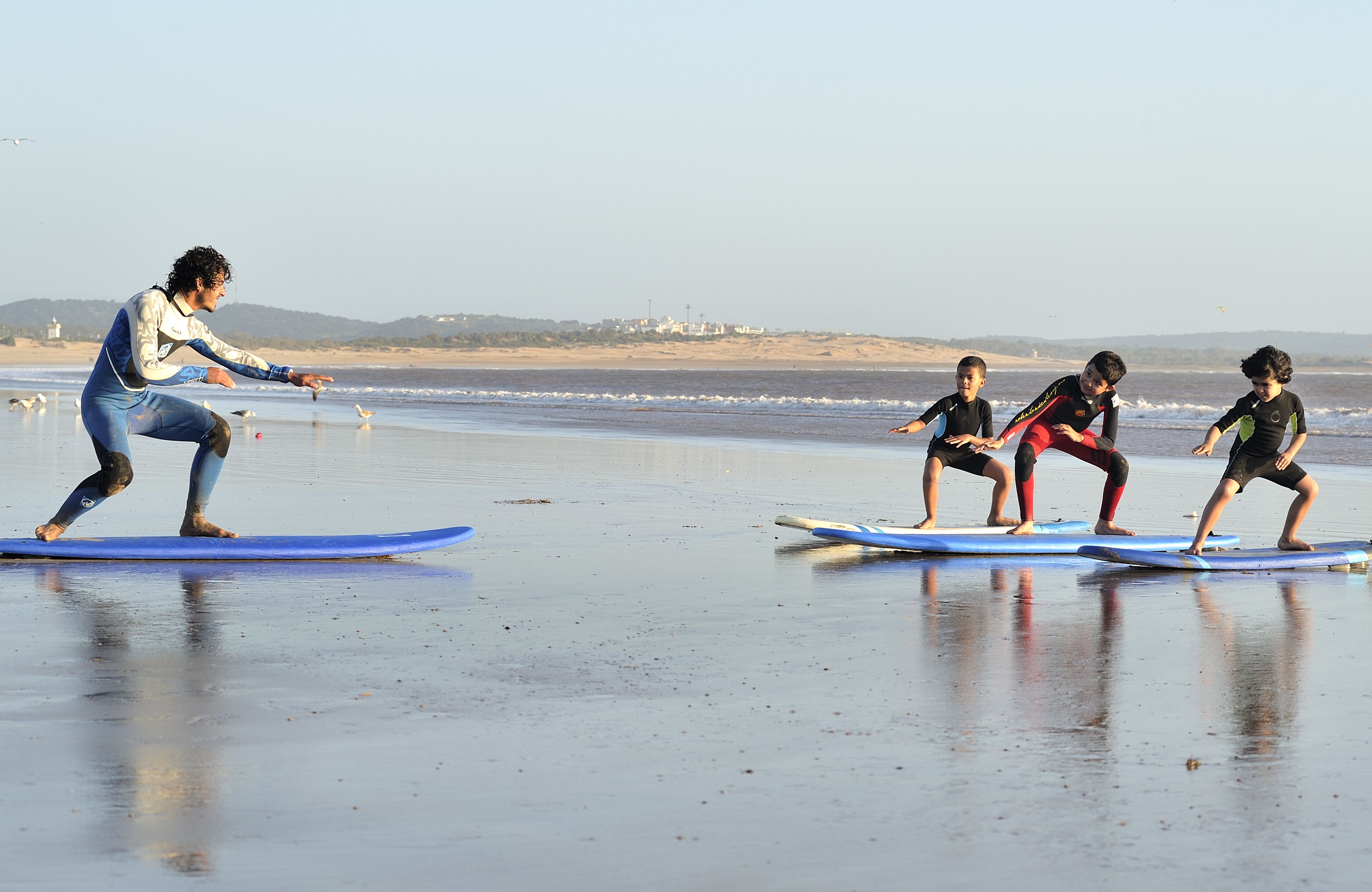 A beach scene with a person teaching surfing to three children on surfboards.