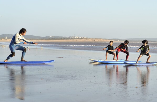 Escena de playa con una persona enseñando a tres niños a surfear.
