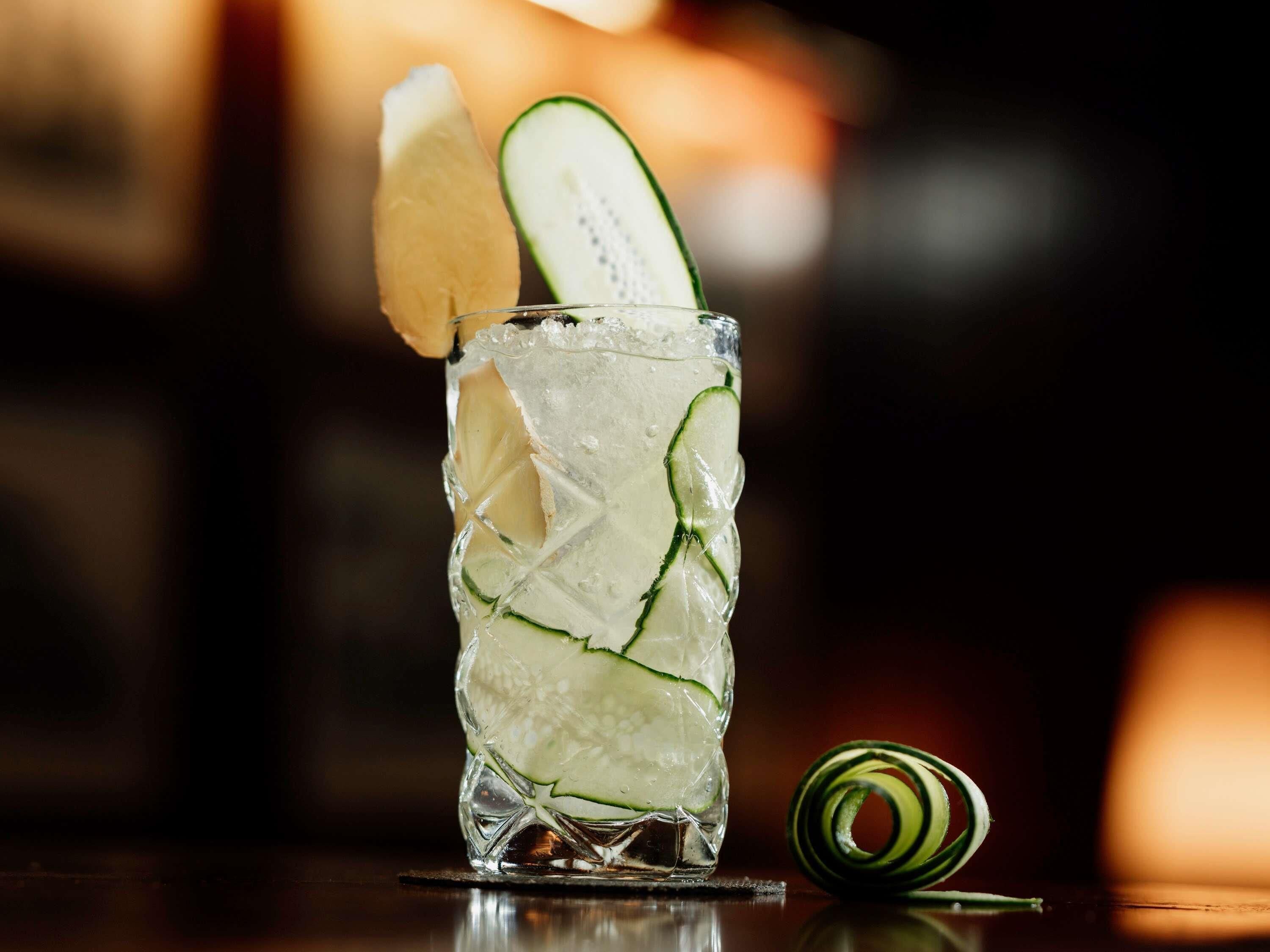 A glass with ice and cucumber slices, a lemon wedge, and a spiral citrus garnish on a reflective surface.