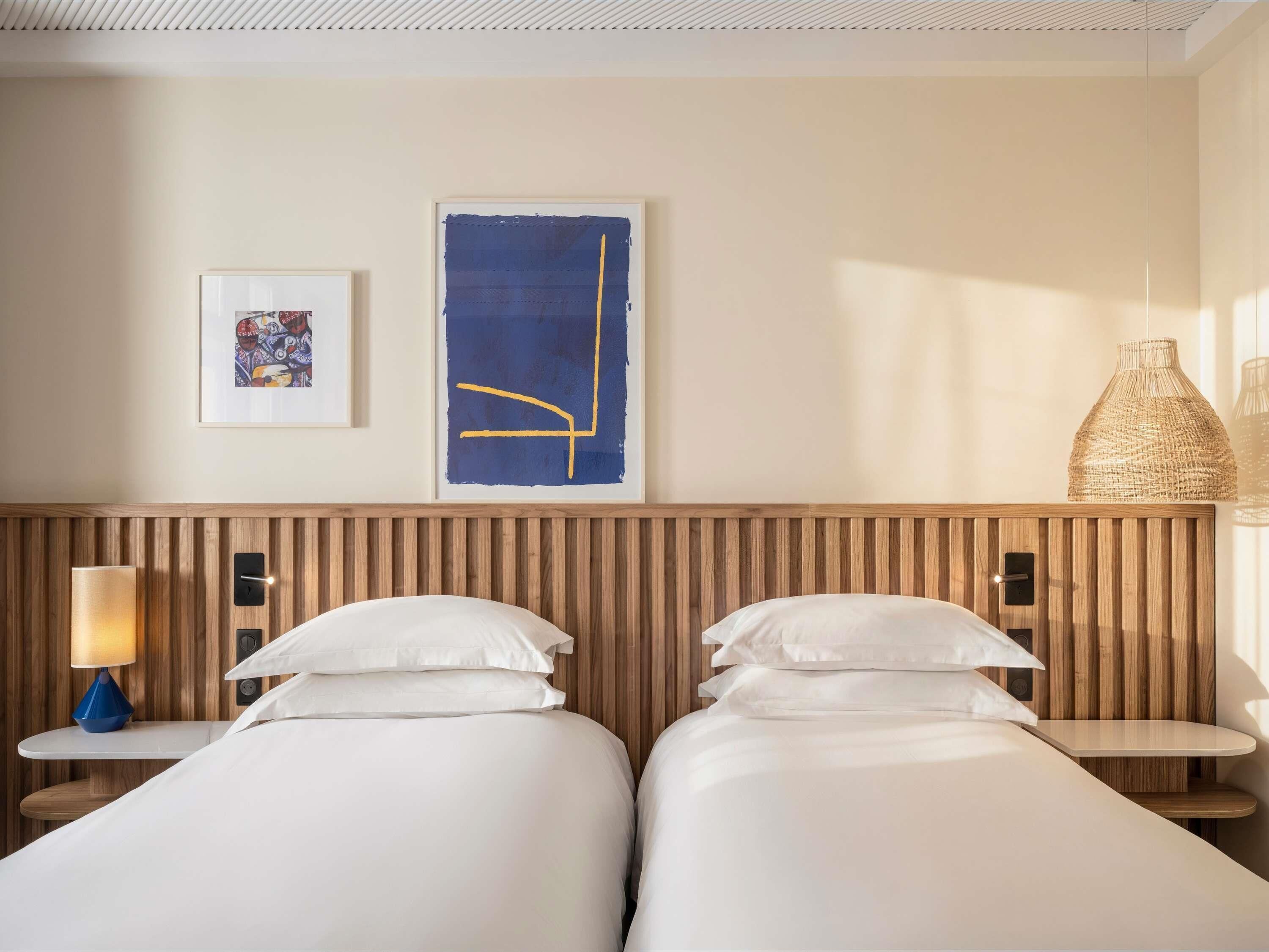 A hotel room with two beds, a wooden headboard, a wall-mounted lamp, and artwork.