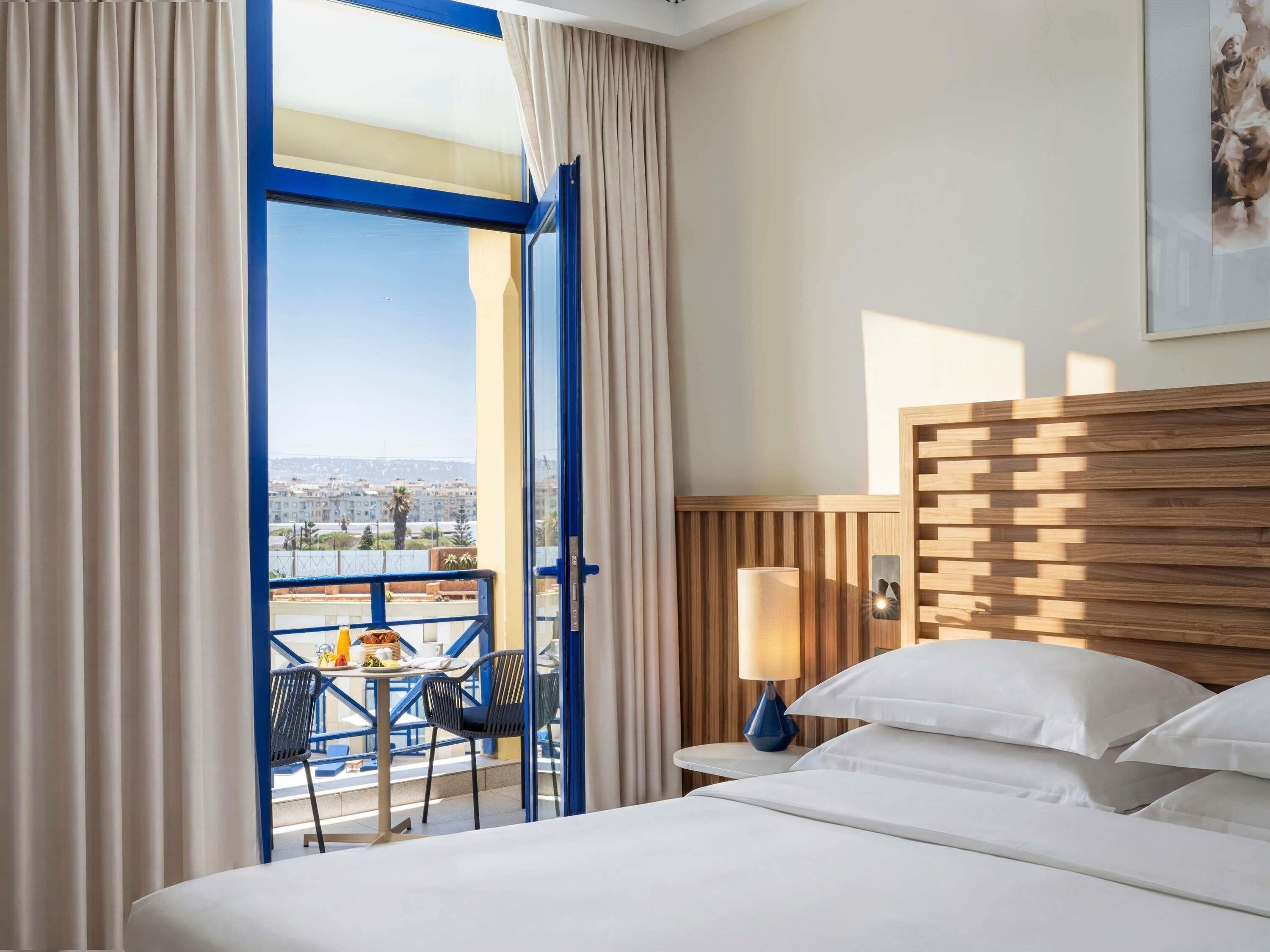 A hotel room with a bed, a nightstand, a lamp, a chair, and a table with a view of the city.