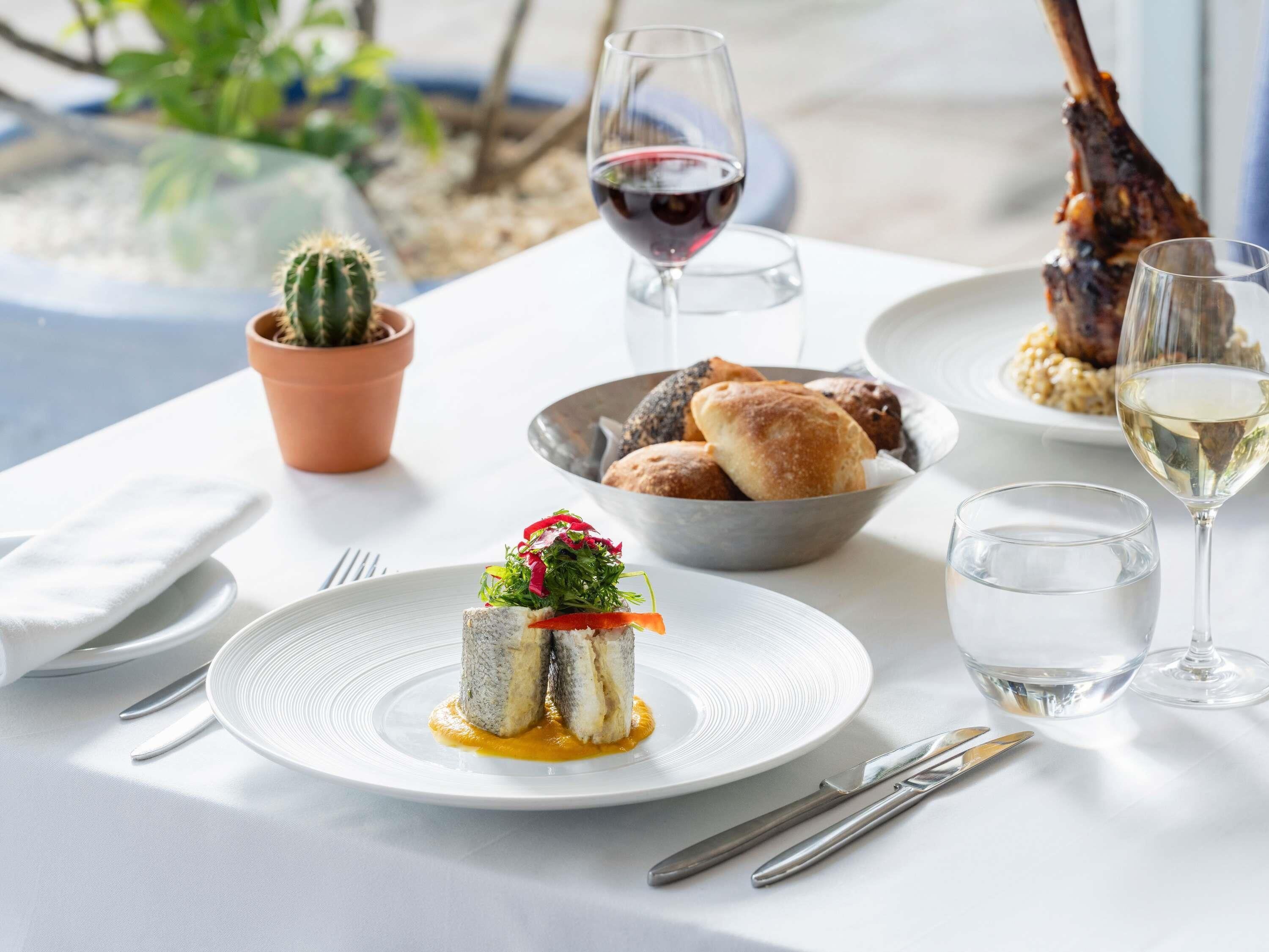Una mesa servida con un plato de pescado, pan, copas de vino y una planta de cactus.
