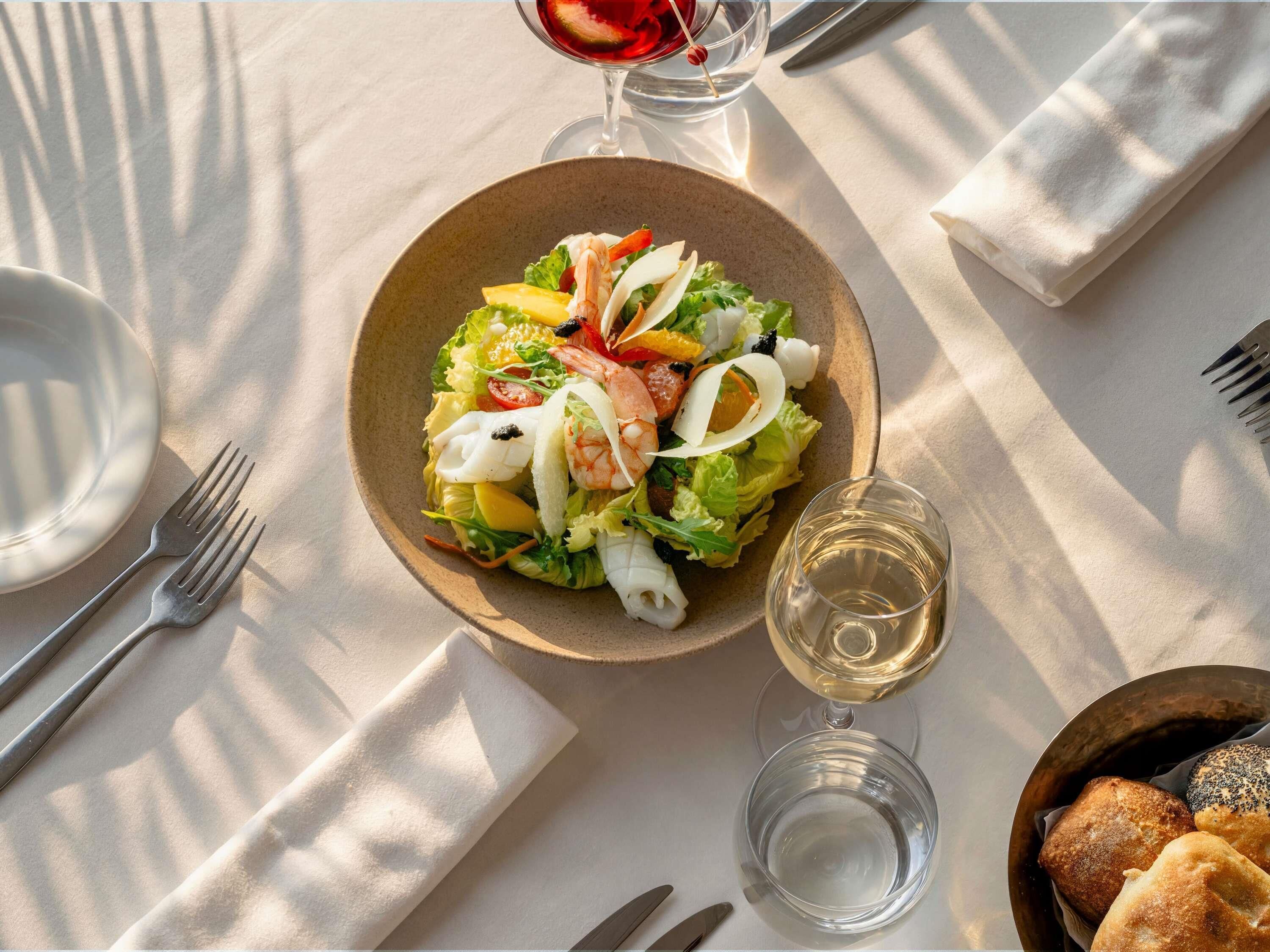 Une table avec une assiette de salade, un verre de vin blanc et un panier de pain.