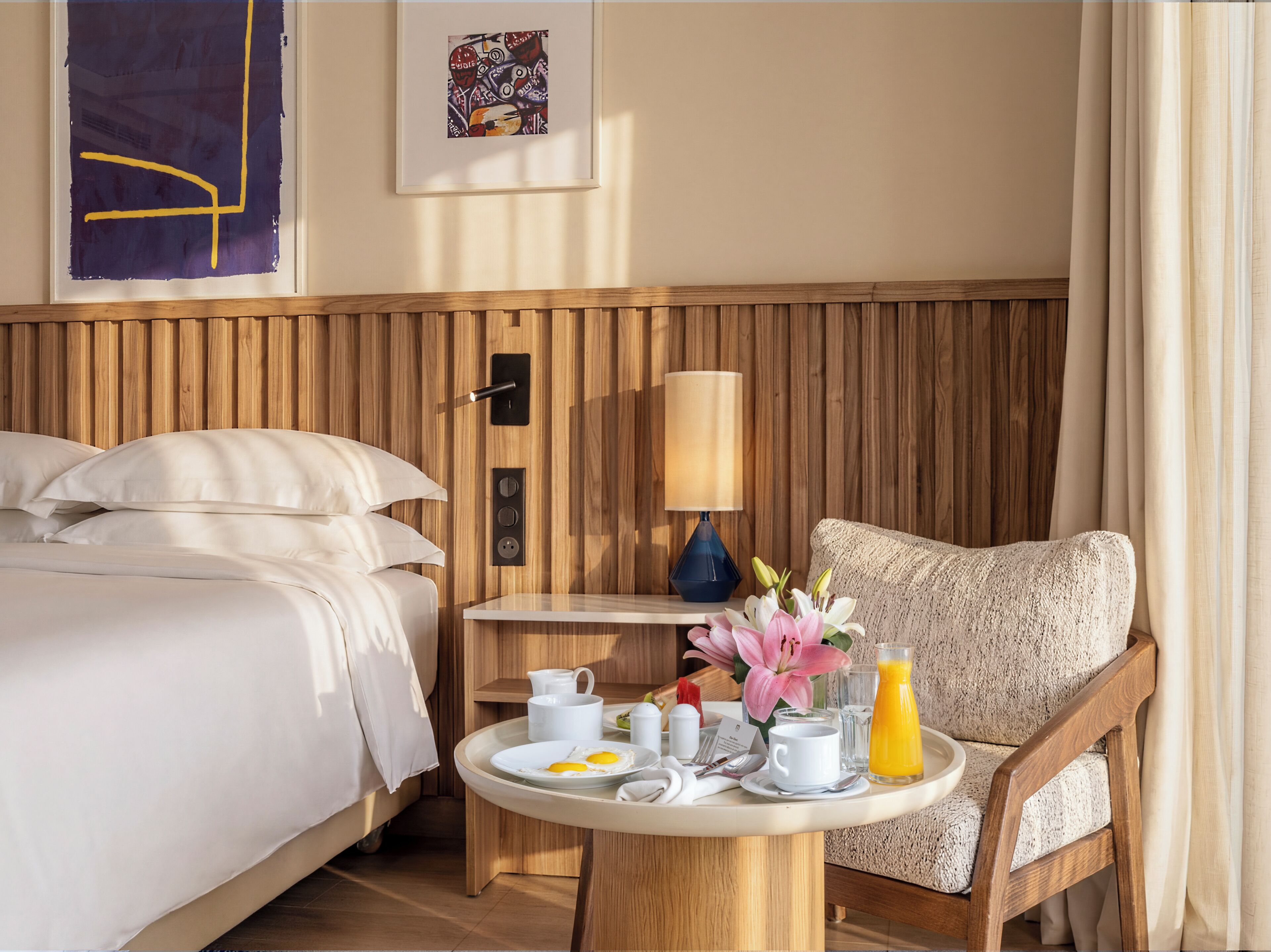 A hotel room with a bed, a chair, a table with breakfast items, a lamp, and a wooden headboard.