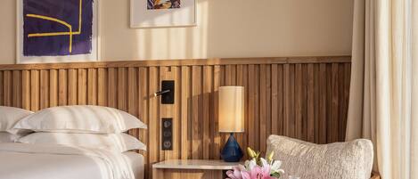 A hotel room with a bed, a chair, a table with breakfast items, a lamp, and a wooden headboard.