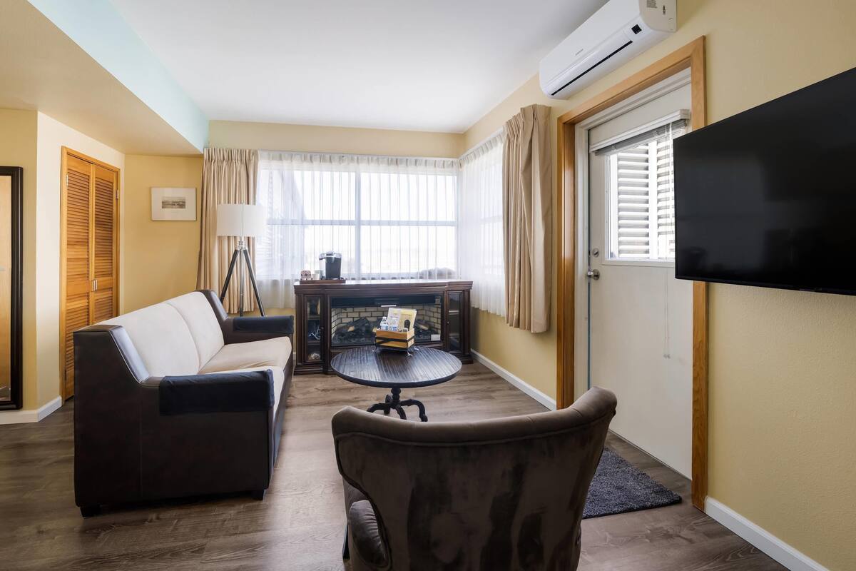 A living room with a sofa, armchair, and a small round table with a lamp. There is a flat-screen TV mounted on the wall and a window with curtains.