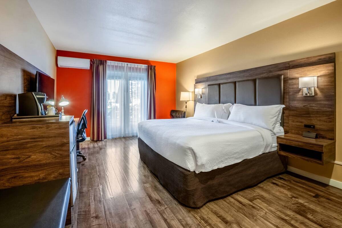 A hotel room with a large bed, wooden flooring, a desk with a chair, a TV, and a window with curtains.