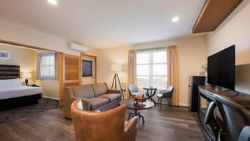 Habitación de hotel con cama, sofá, sillón, mesa de comedor, sillas, televisor y un refrigerador pequeño.
