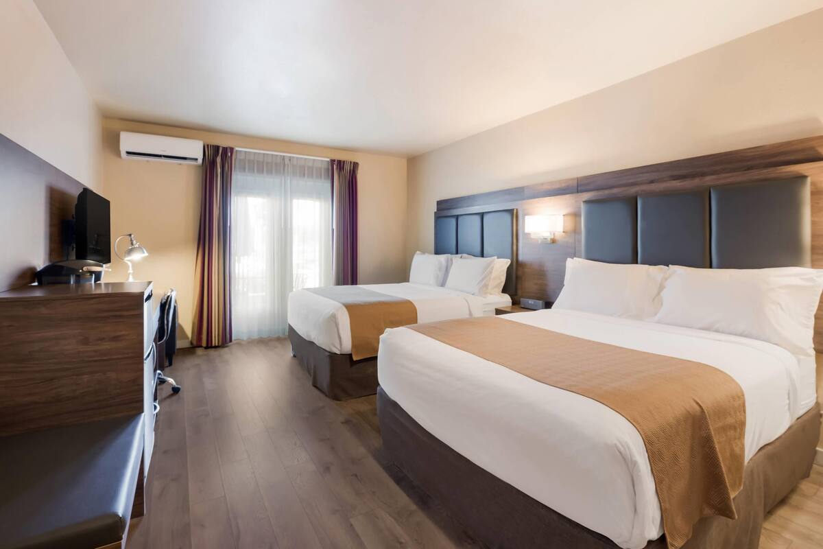 A hotel room with two beds, a desk, a TV, and an air conditioning unit.