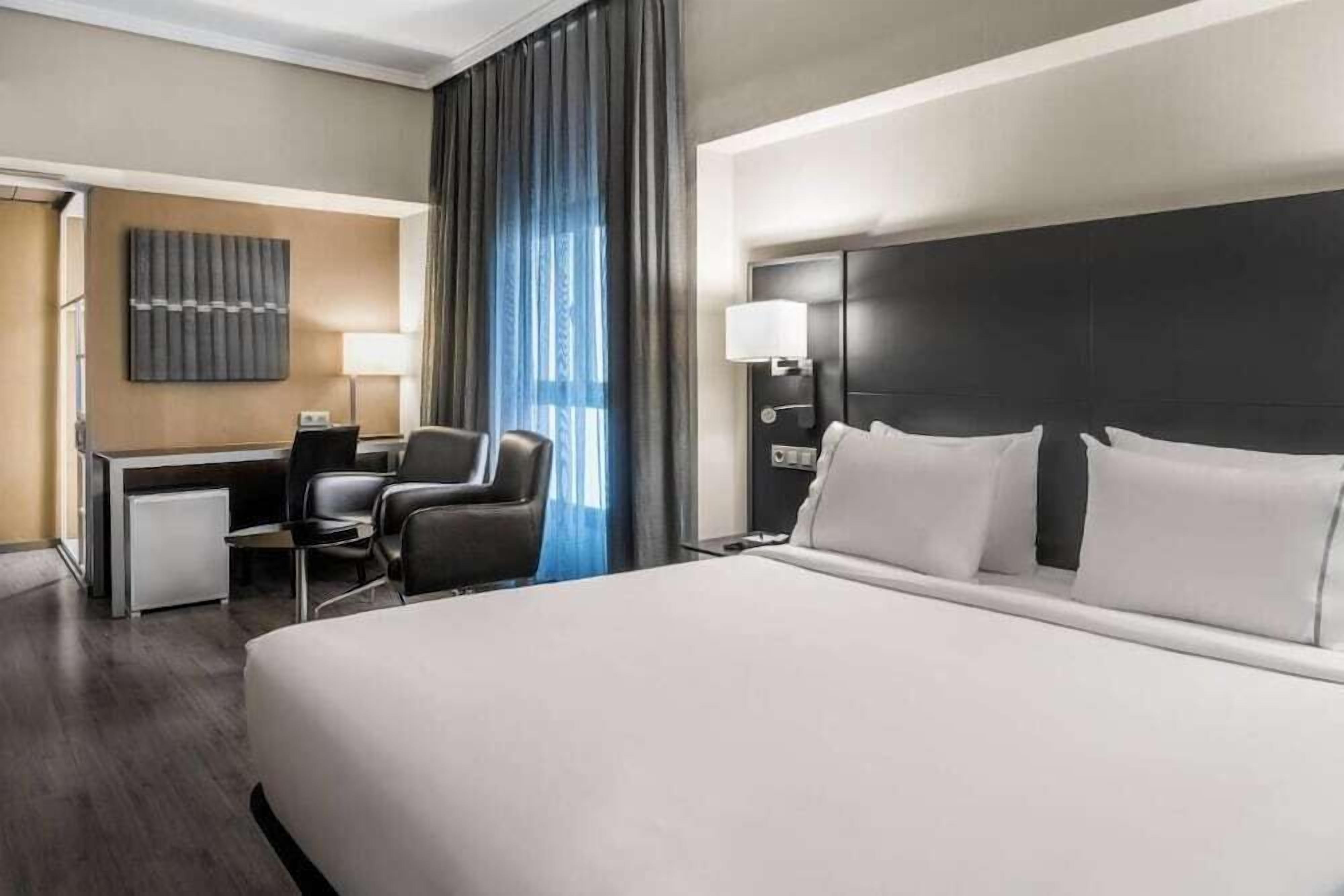 Habitació d'hotel amb un llit gran, una escriptori amb cadira, un frigorífic petit i una làmpada de paret.