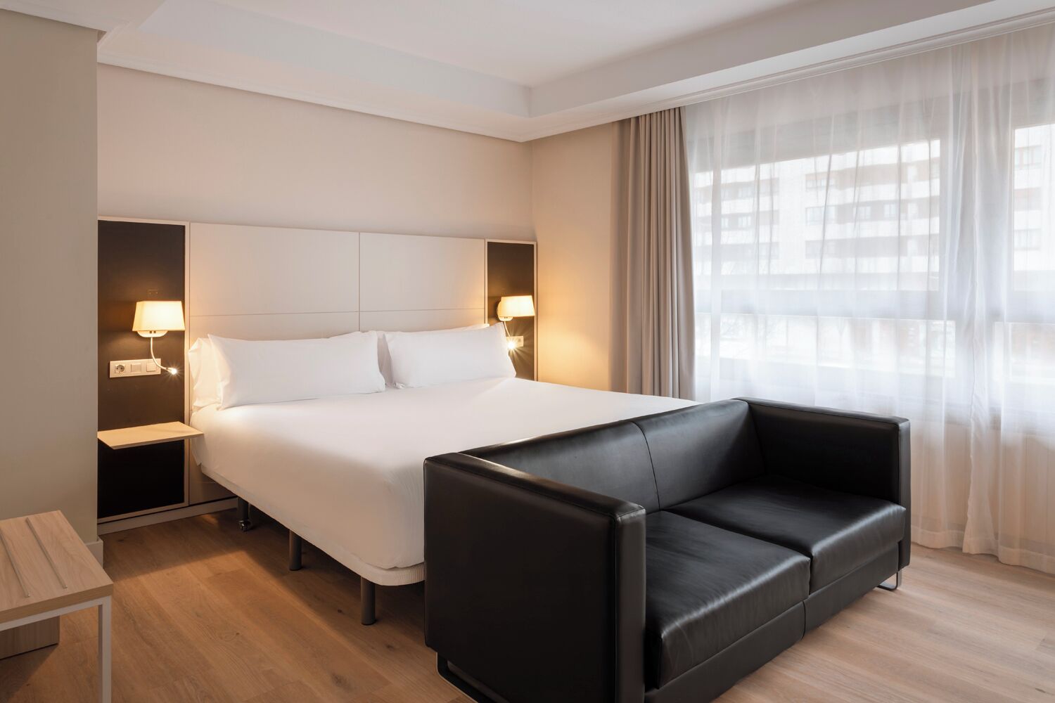 Habitació d'hotel amb un llit, dues taules de nit amb làmpades, un sofà de pell negra i una finestra amb cortines translúcides.