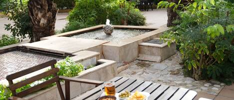 Un área de comedor al aire libre con una mesa preparada para dos, una pequeña fuente y diversas plantas y árboles al fondo.