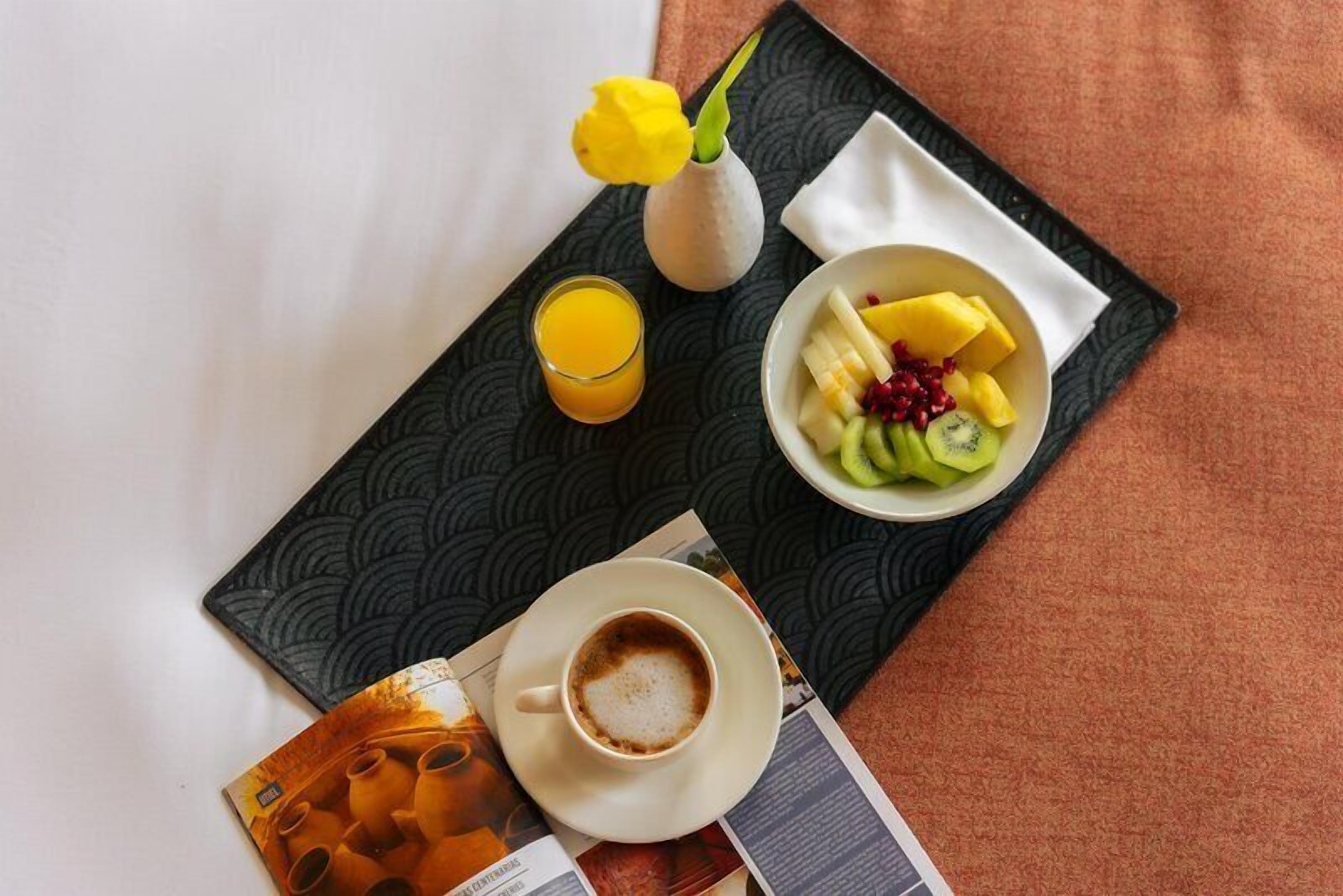 Un plateau de petit-déjeuner avec une tasse de café, un verre de jus d’orange, un bol de fruits et un vase contenant une fleur jaune.