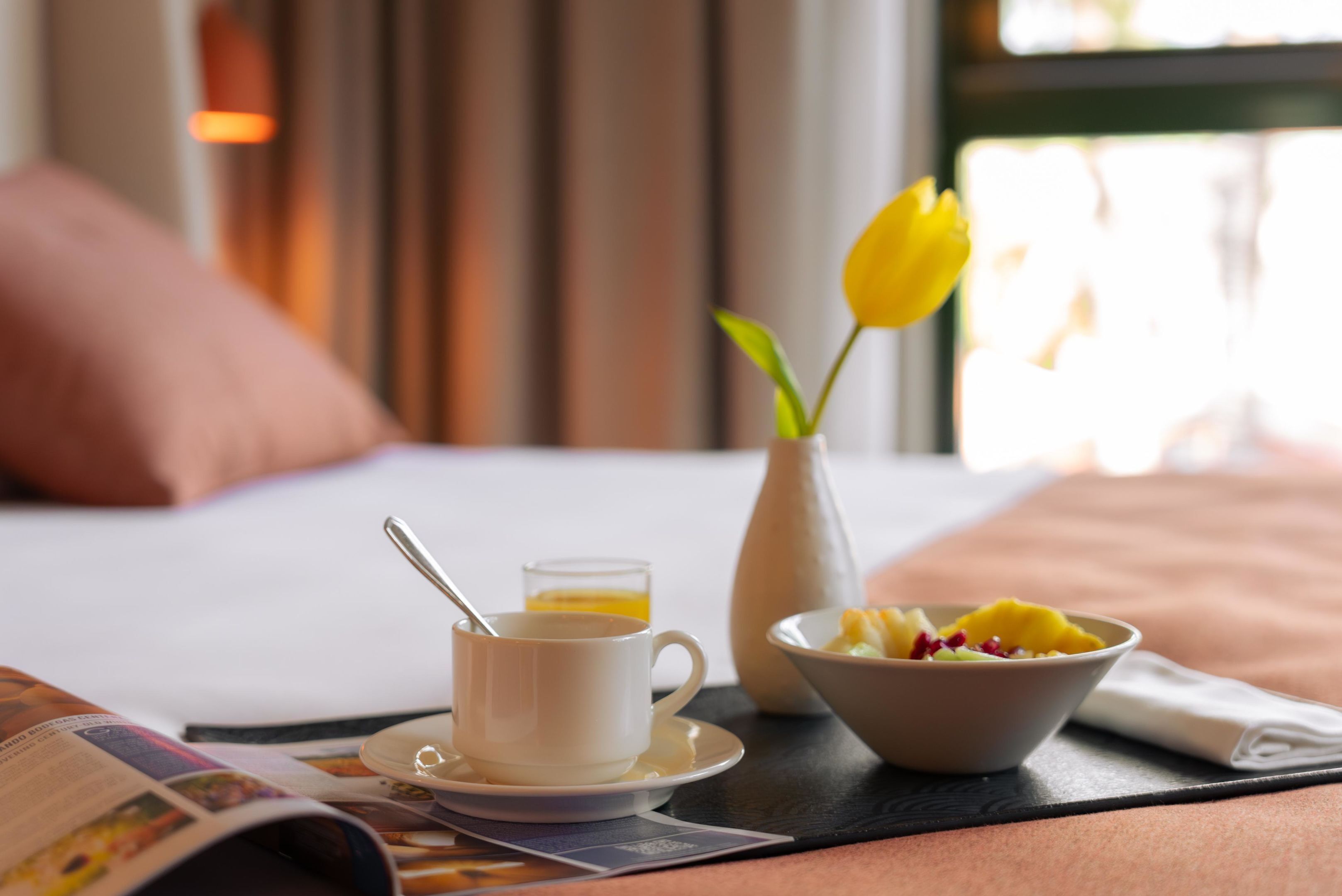A breakfast setting with a cup of coffee, a bowl of fruit, and a vase with a single yellow flower on a table.
